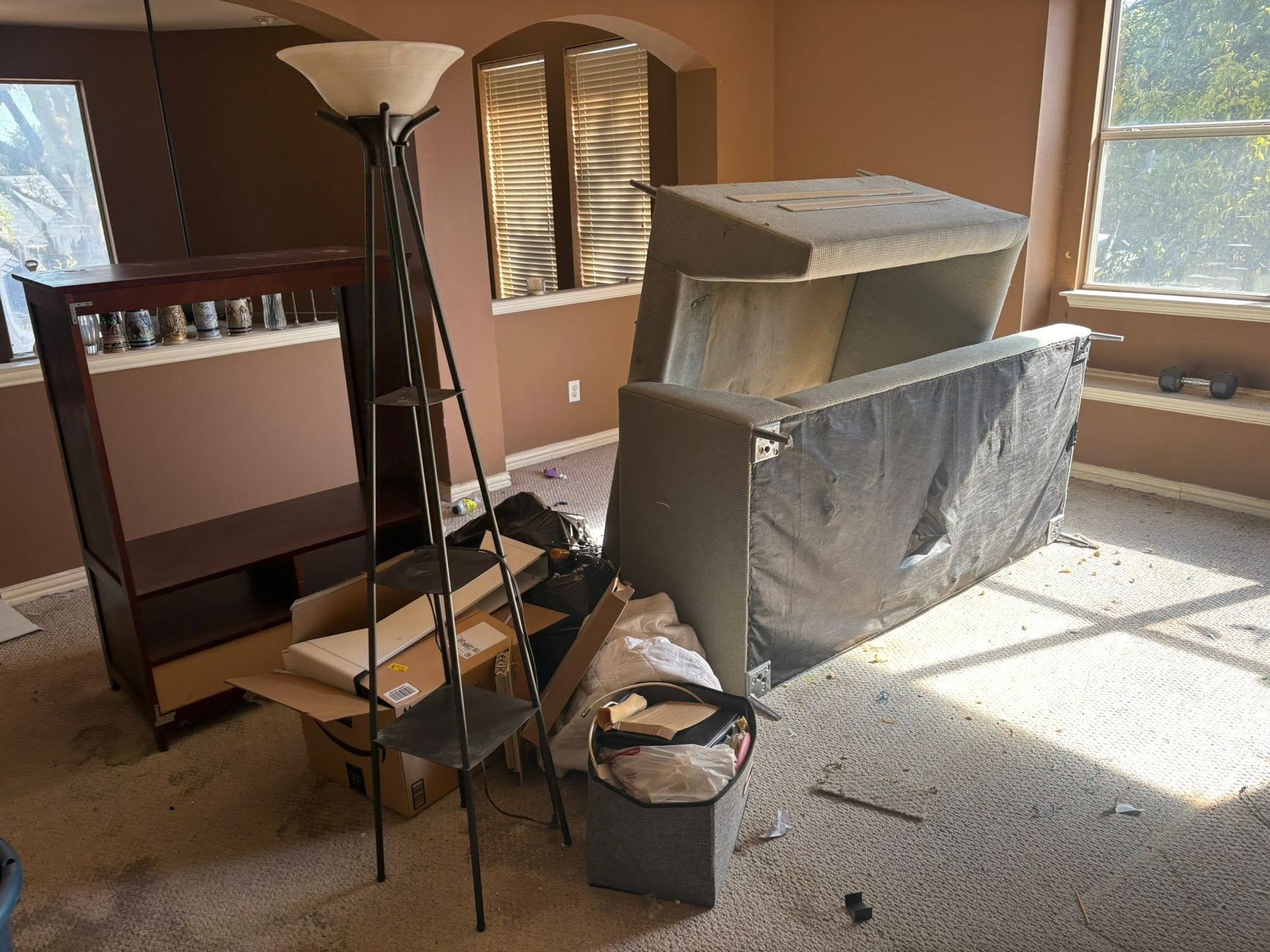 Disassembled furniture and boxes in a sunlit room with windows and a floor lamp.