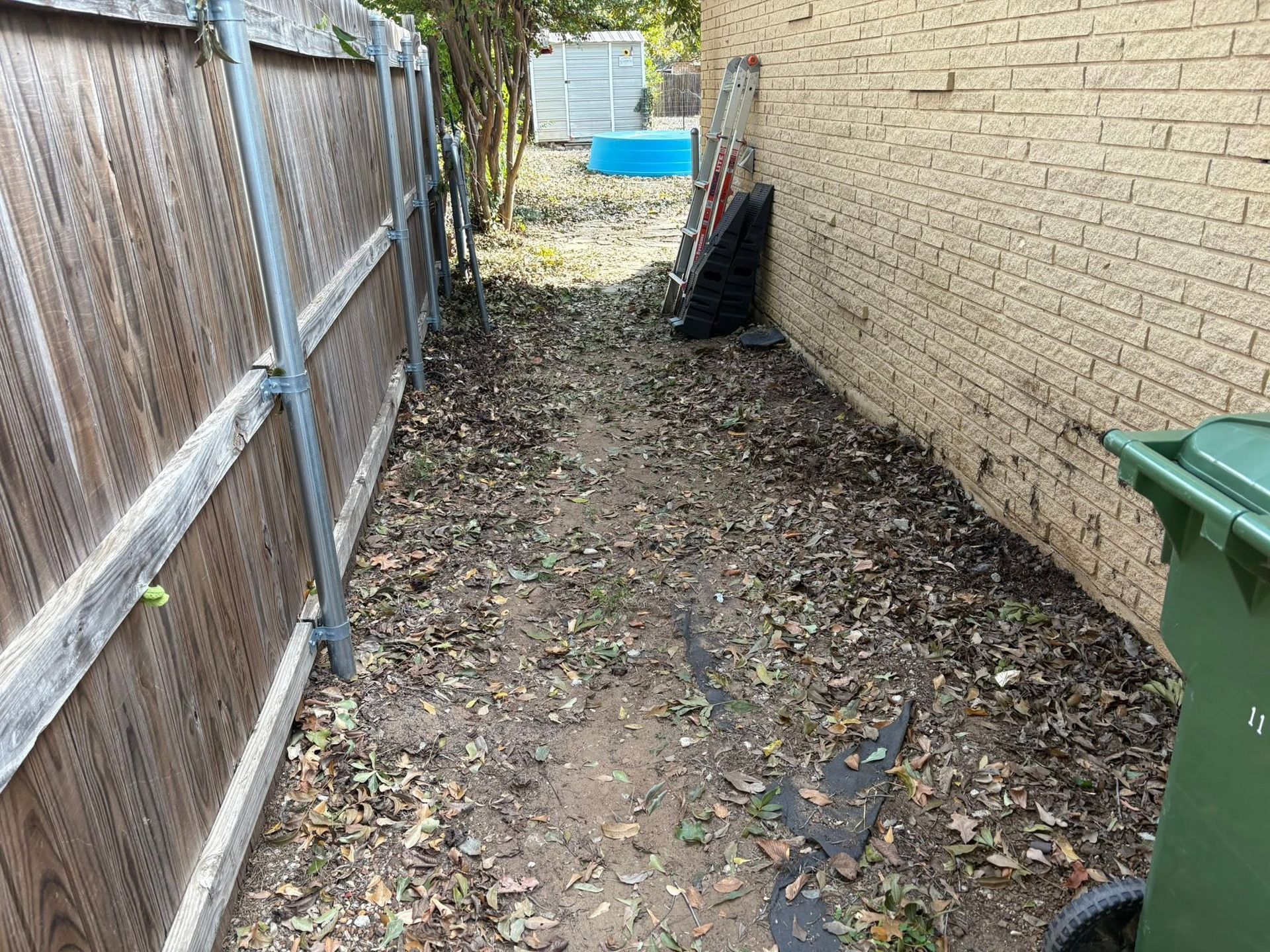 Narrow muddy side yard between a wooden fence and brick wall, with trash bins and blue pool in the distance