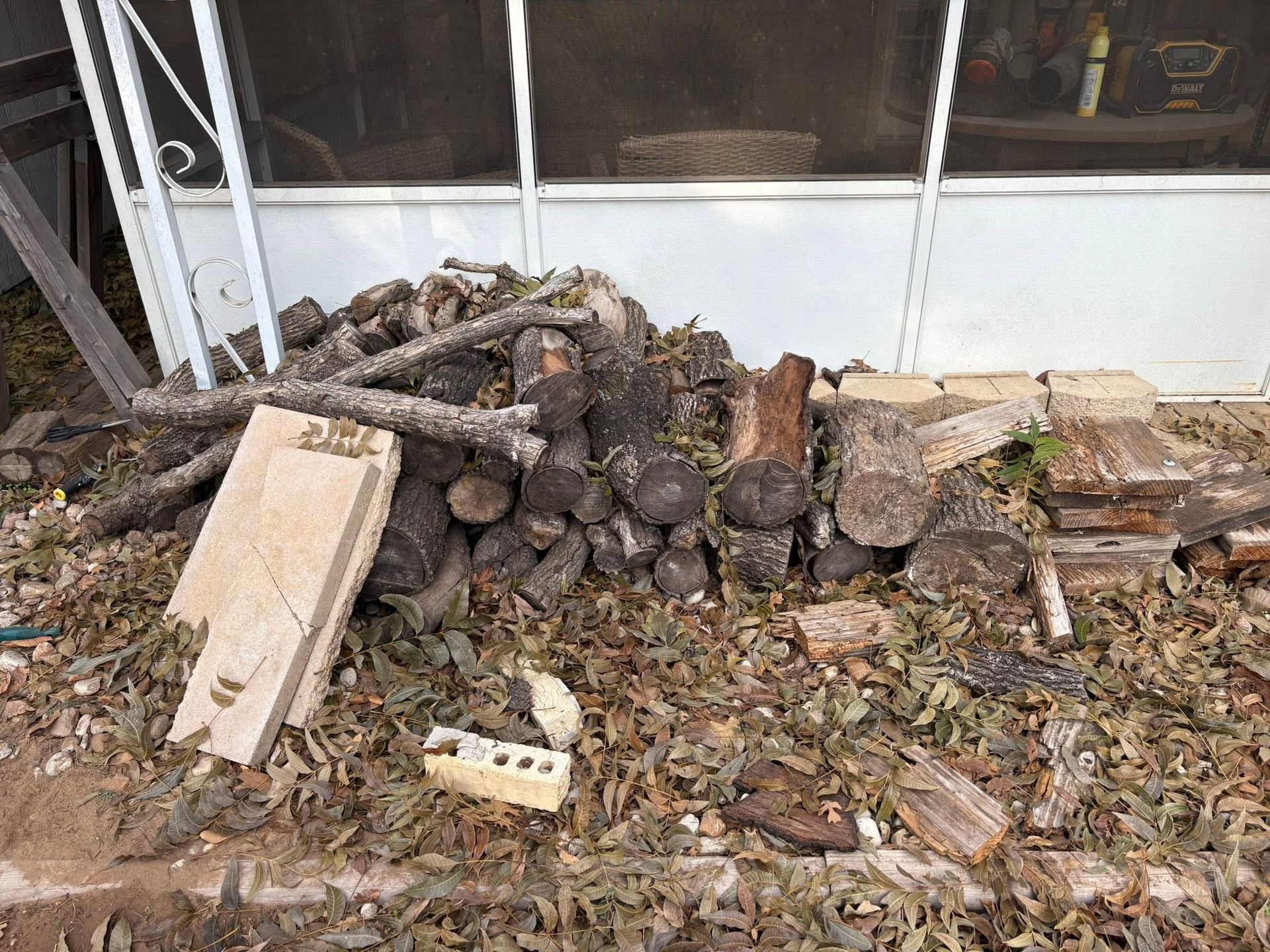 Pile of firewood and debris beside a white house wall, with a board and bricks on the ground.