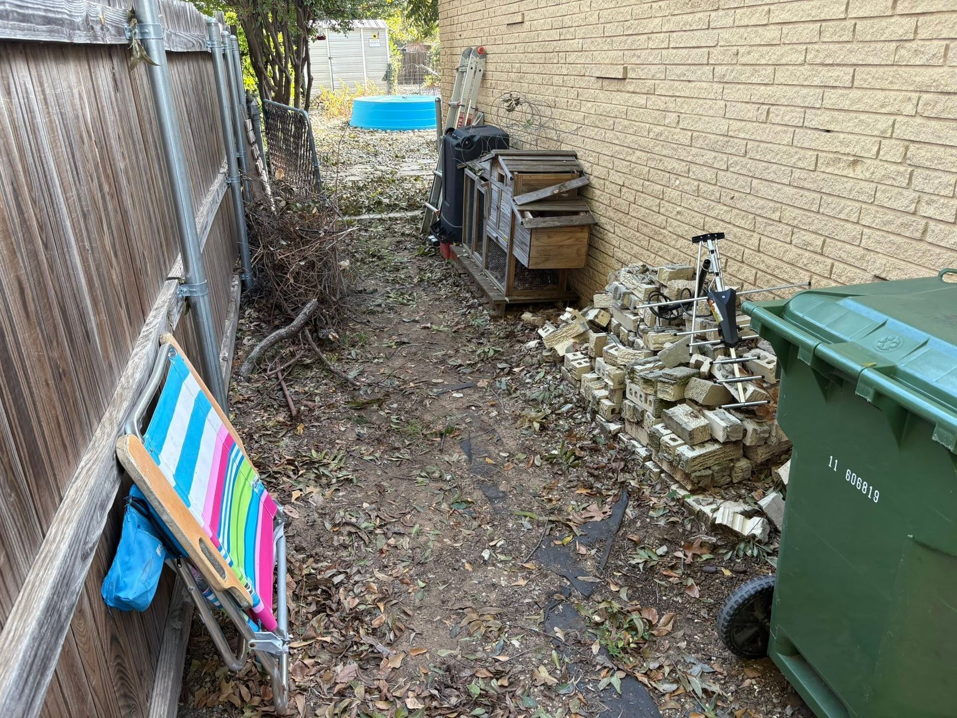 Narrow side yard with debris, stacked wood, green trash bin, and colorful boards leaning against a fence.