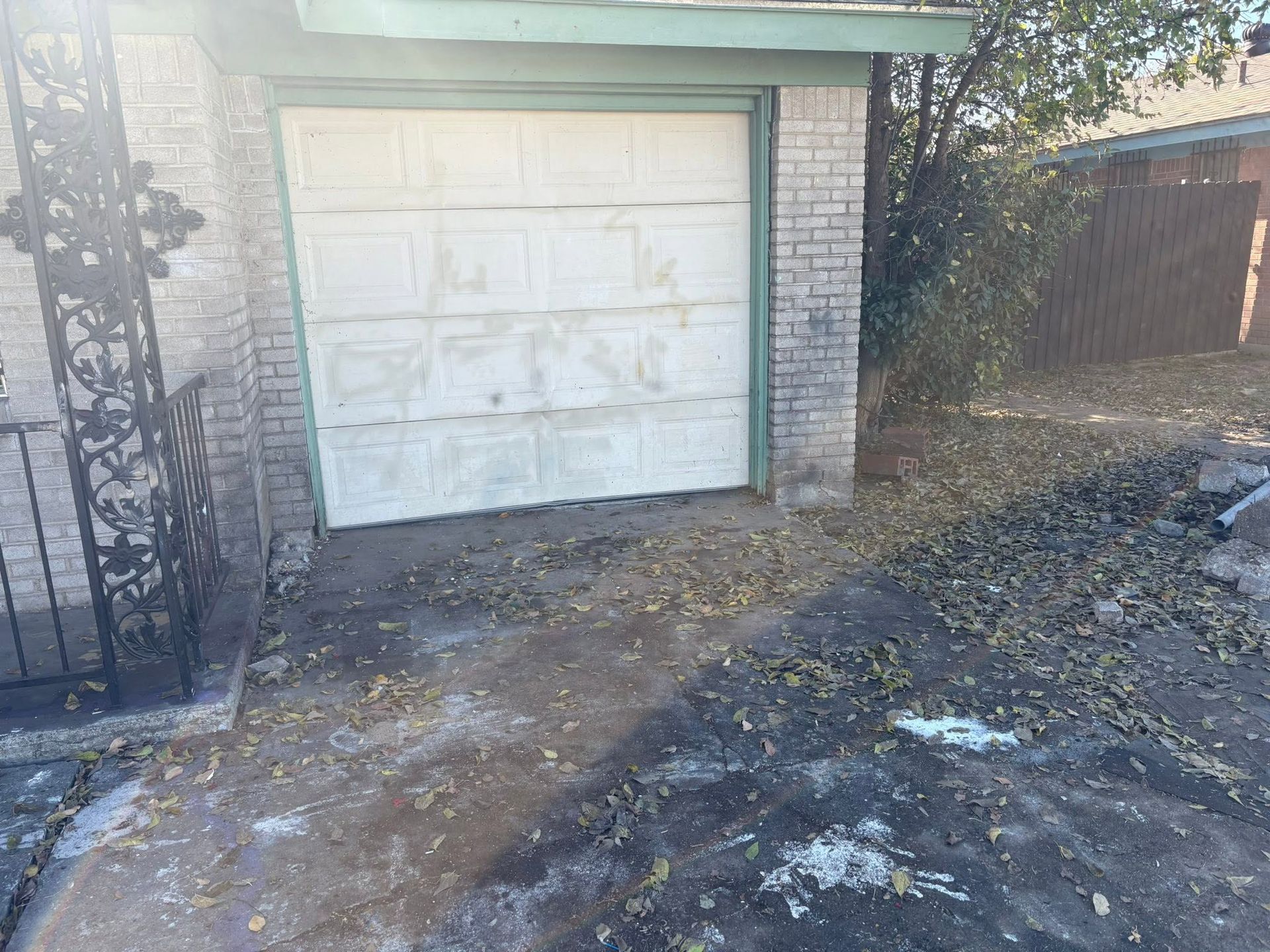 White garage door beside a muddy yard and fence, with a brick wall and scattered debris.