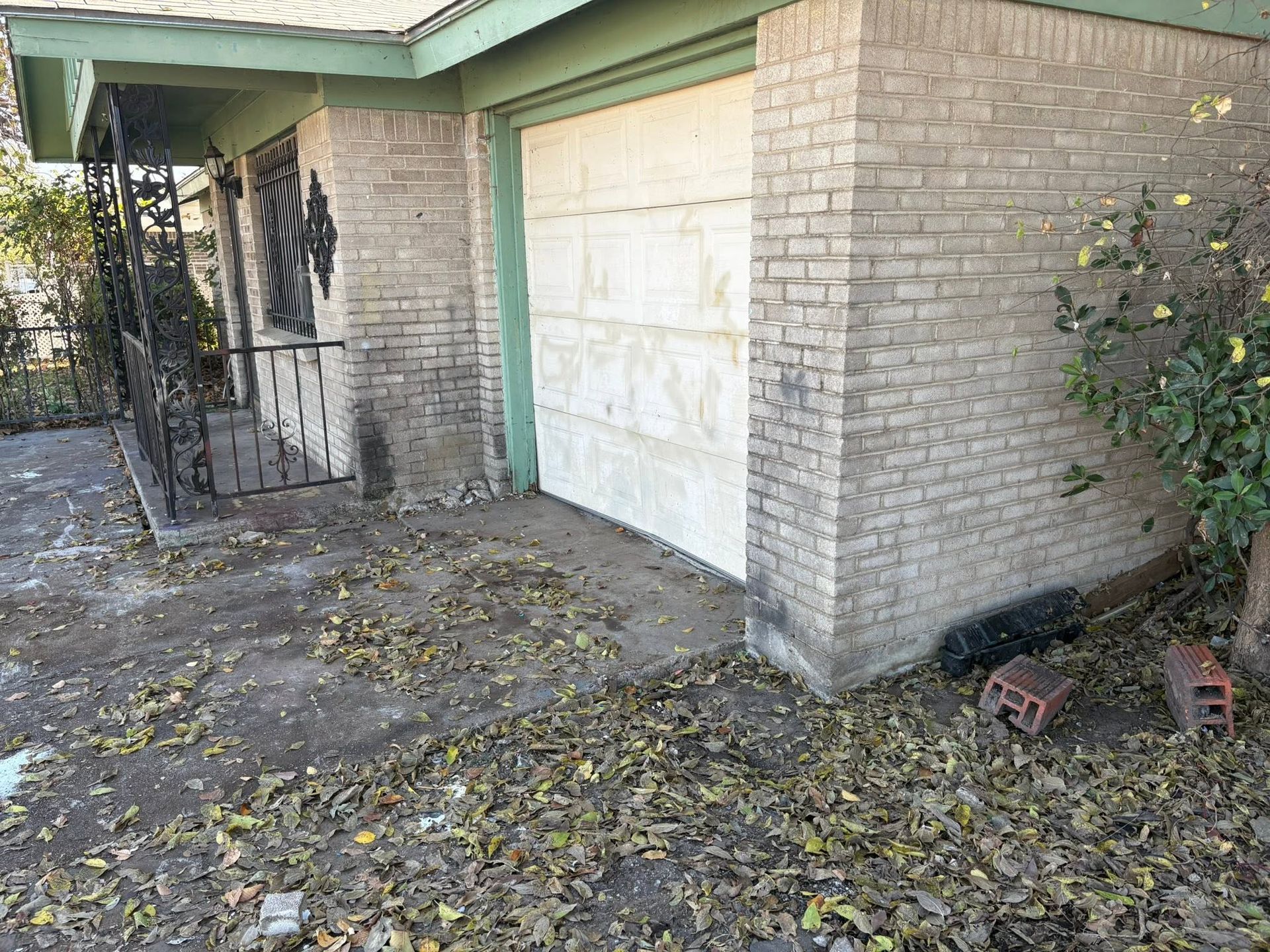 Brick house side entrance with a white garage door, overgrown yard, and scattered leaves