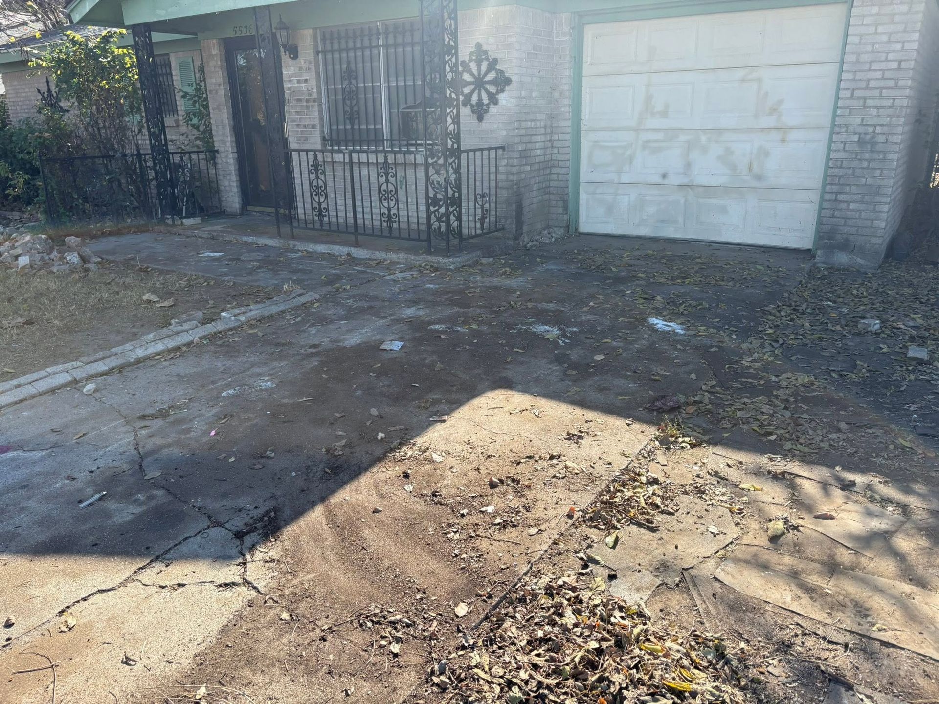 Sunlit driveway with cracked pavement, a metal gate, and a white garage door in front of a brick building
