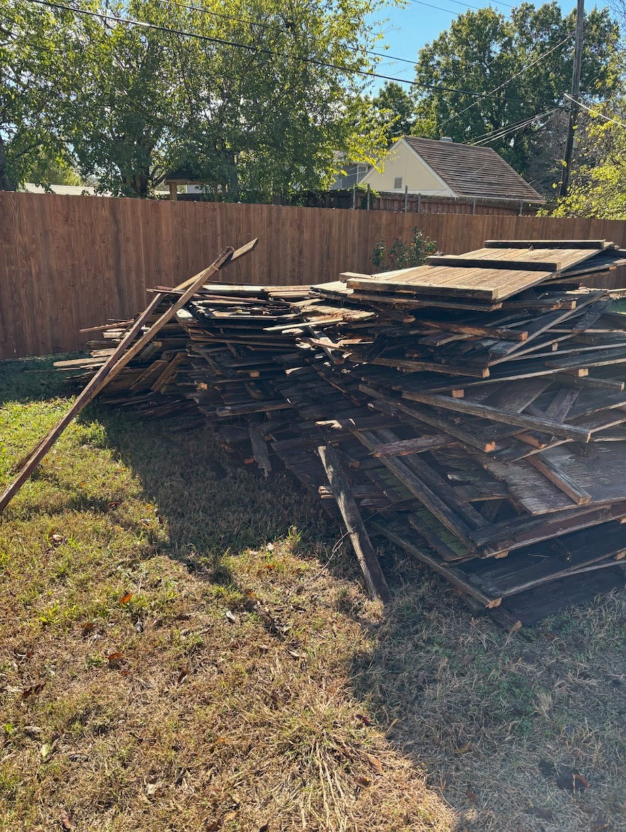 Stacked weathered wood planks in a backyard beside a wooden fence on a sunny day