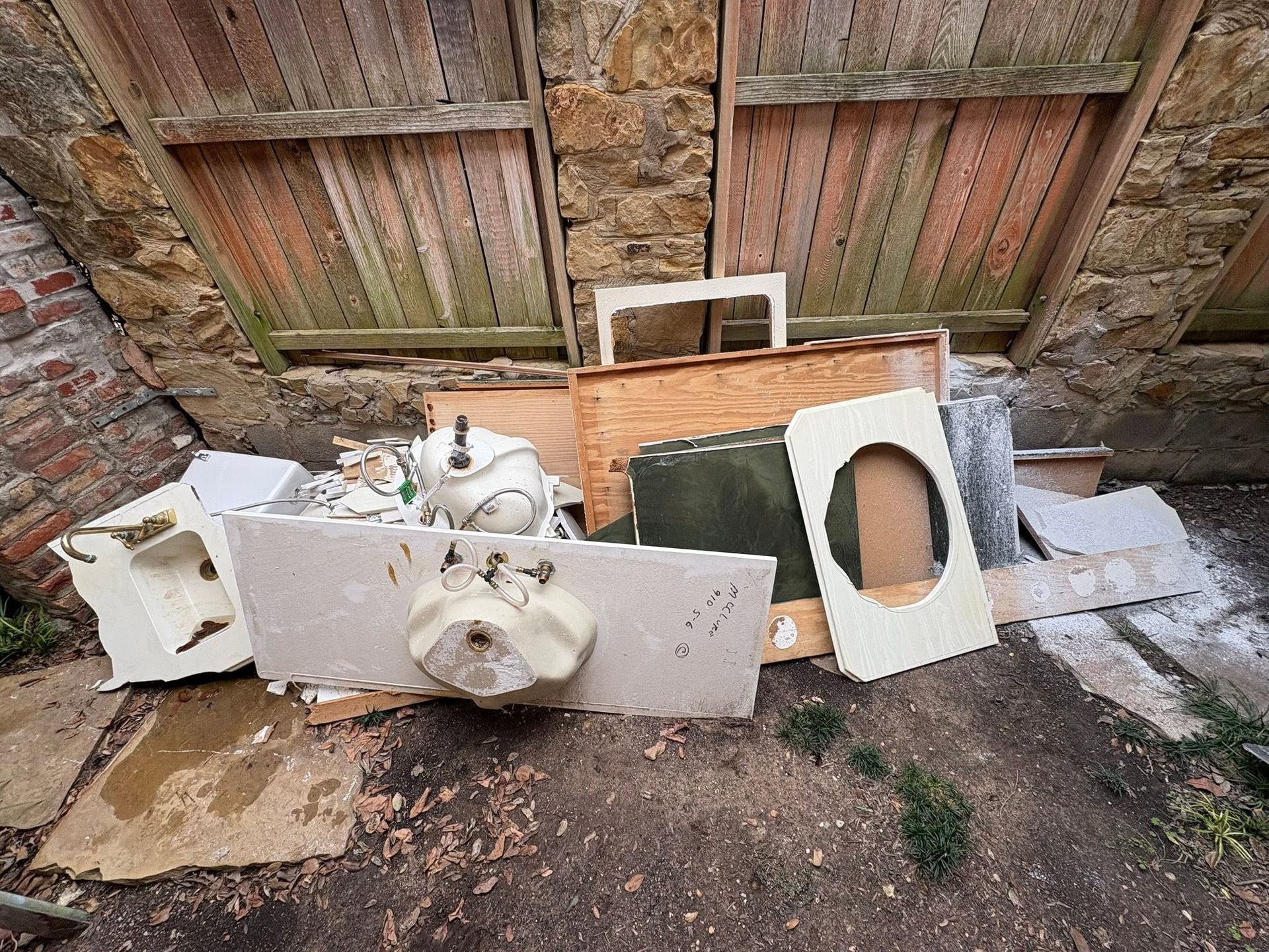 Discarded sink and broken furniture pieces piled against a wooden wall outdoors.