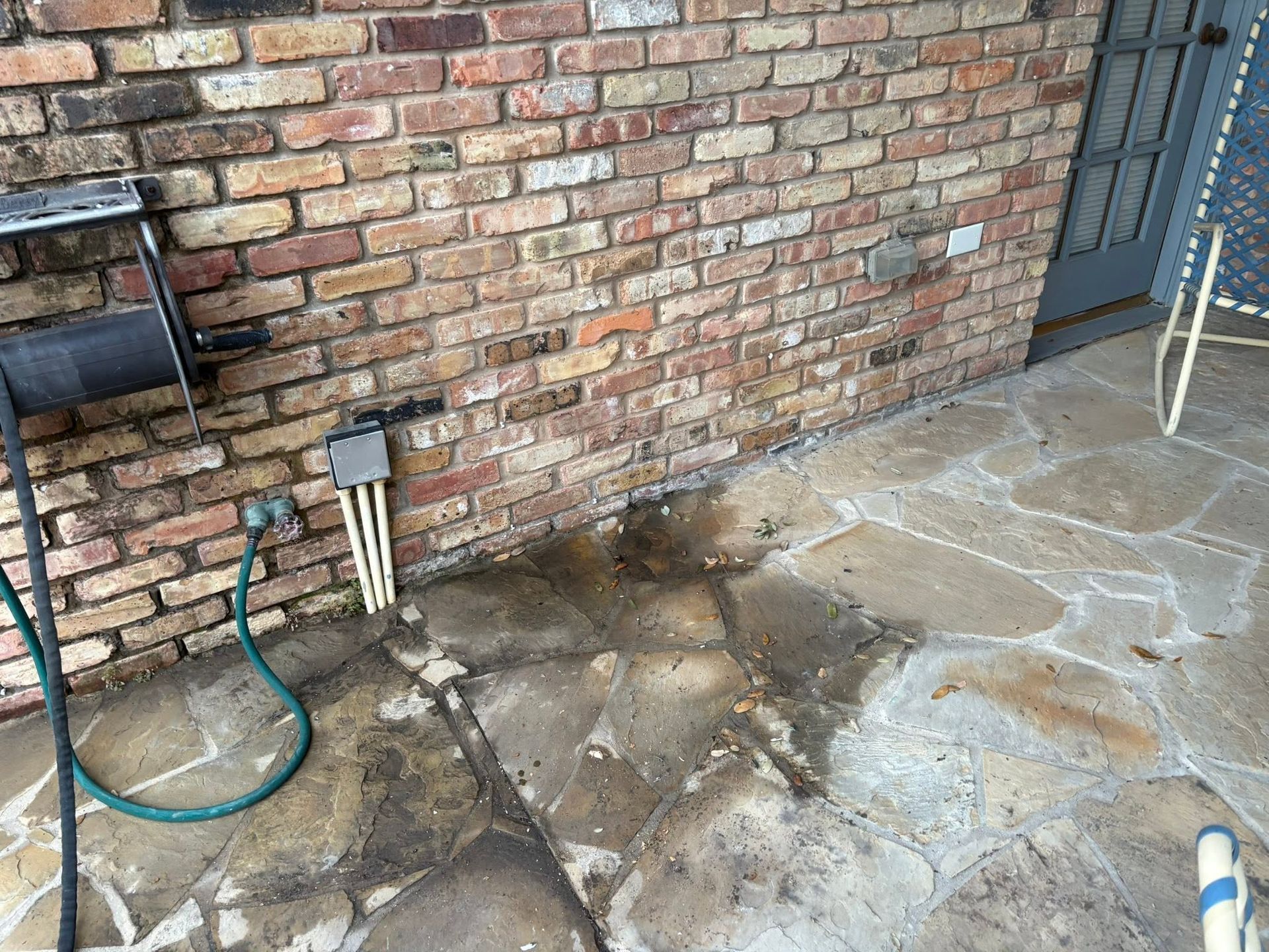 Brick wall patio with wet concrete stains, a hose, and utility pipes near the ground