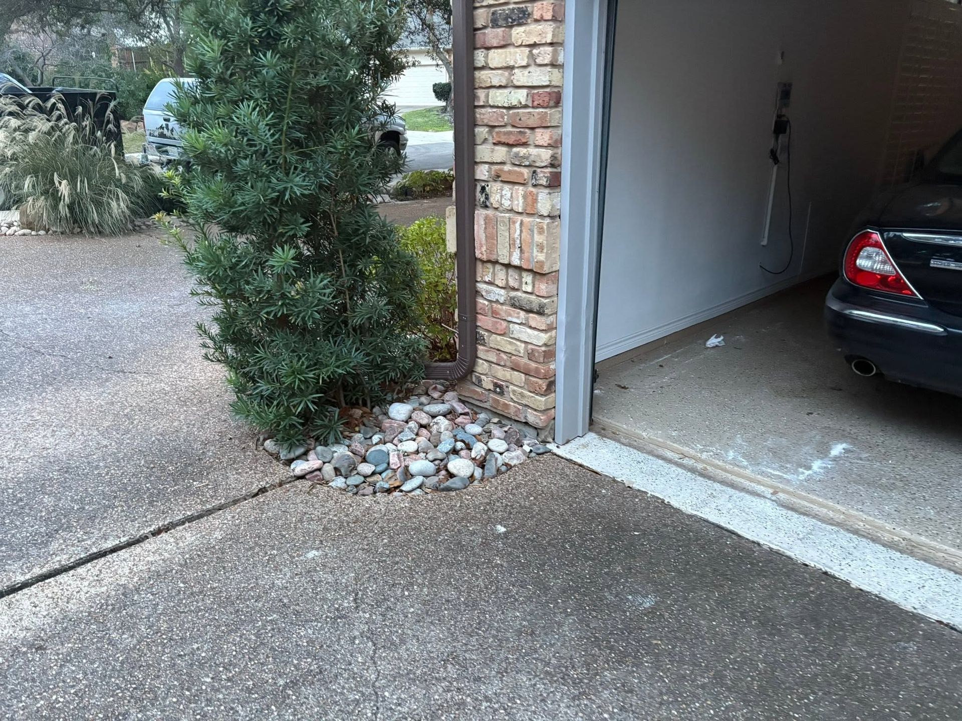 Driveway leading to an open garage with a black car parked inside and a brick wall with shrubs nearby