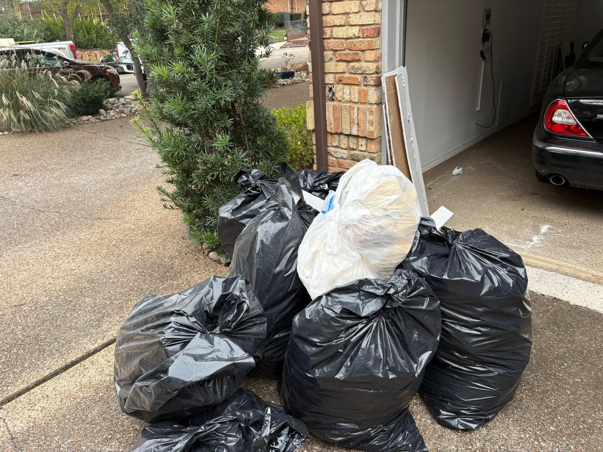 Pile of black trash bags and a white bag stacked beside a garage driveway.