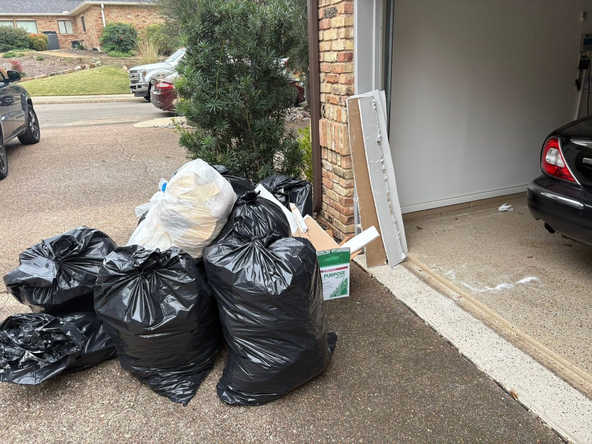 Several trash bags piled beside a garage in a driveway, with a parked car inside.