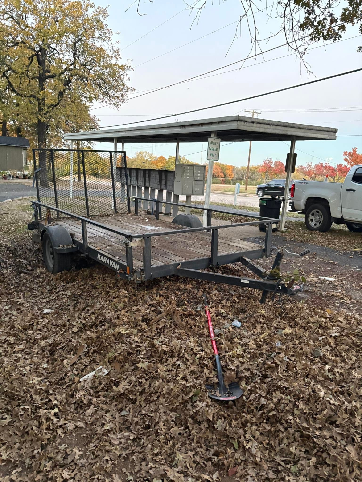 Open utility trailer under trees, covered in fallen leaves, with a truck parked nearby.