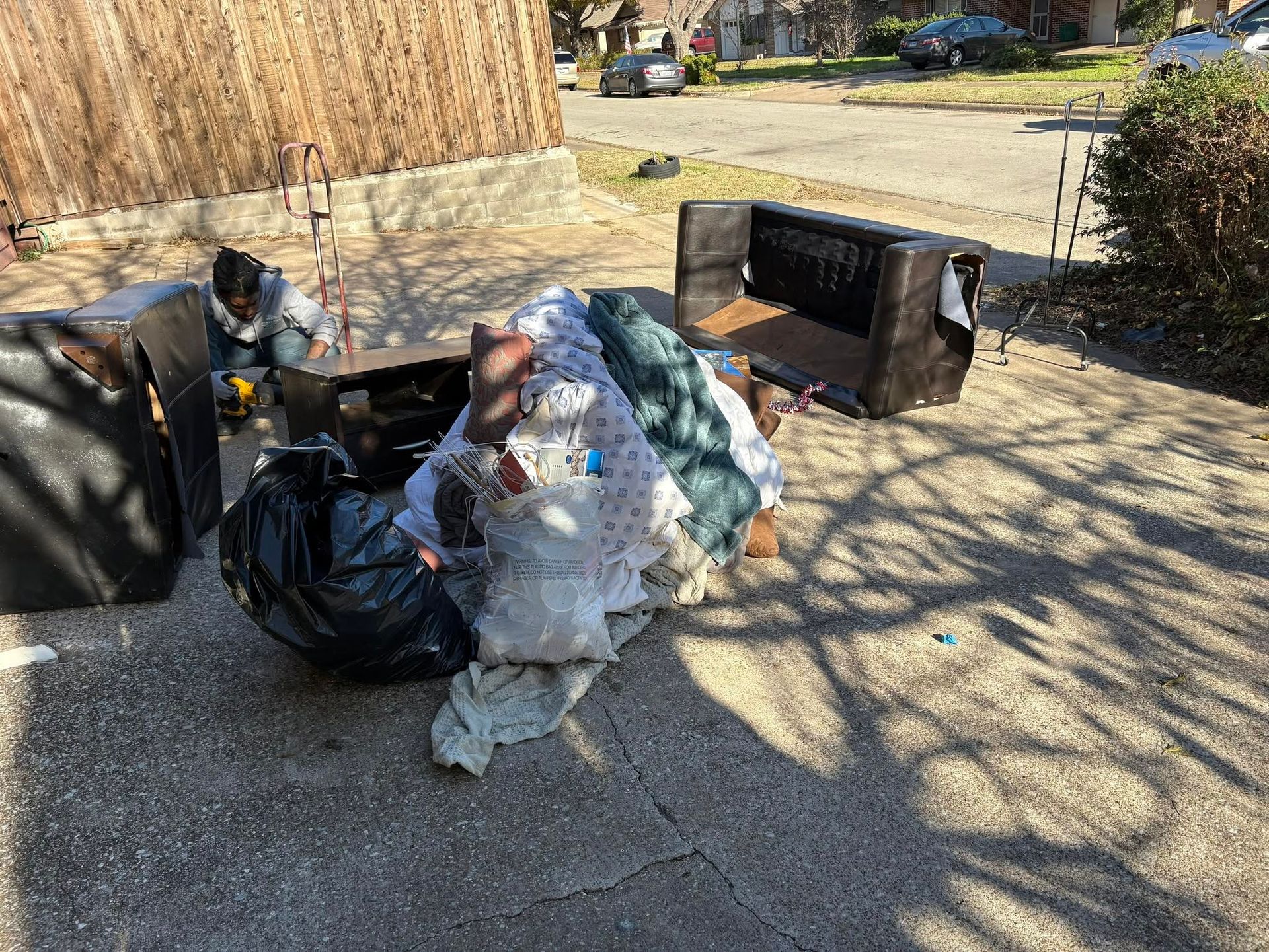 Piles of trash bags and discarded items scattered on a driveway beside an open black dumpster.