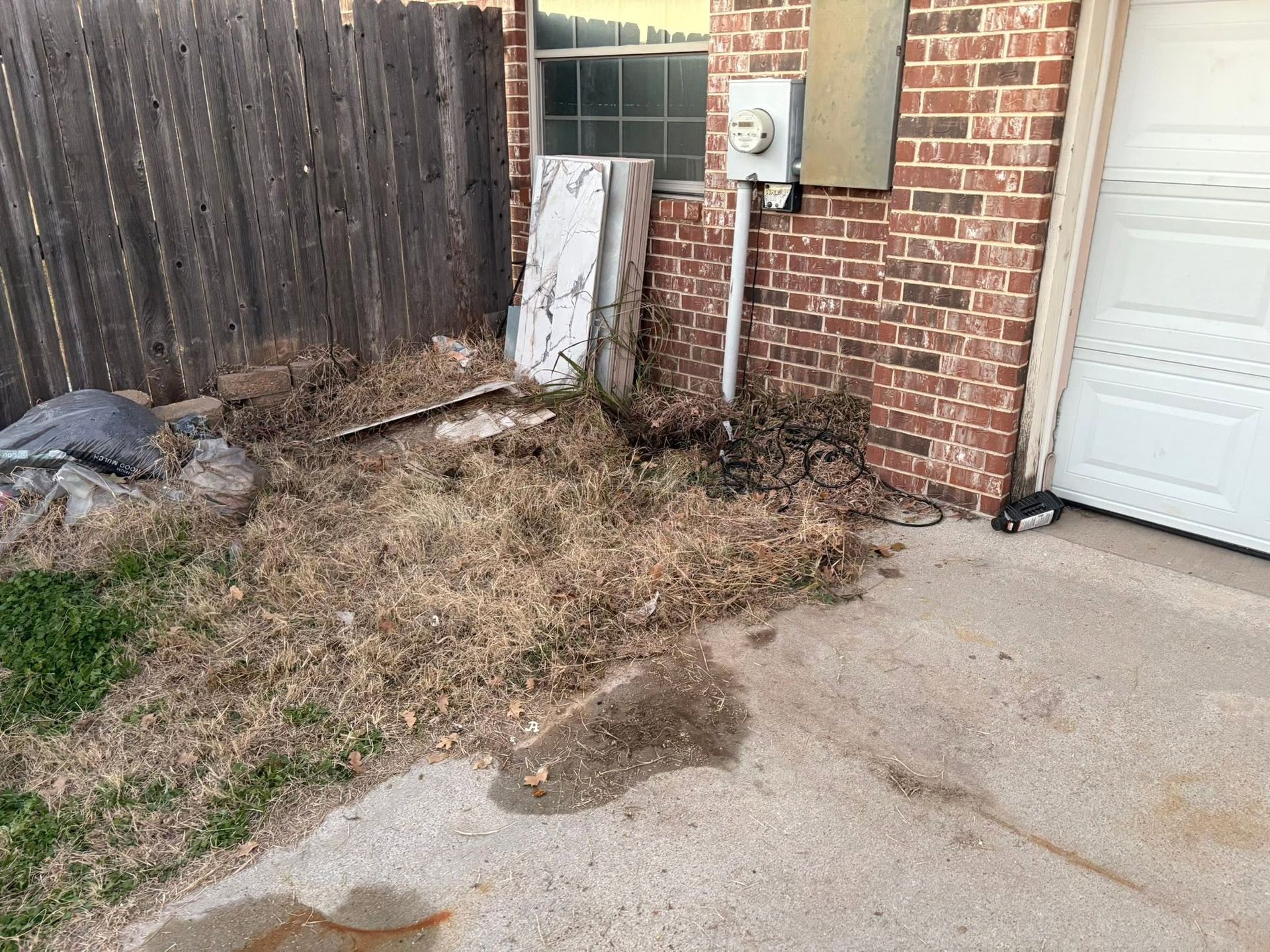Overgrown weeds and dead grass beside a brick house and driveway near a white garage door
