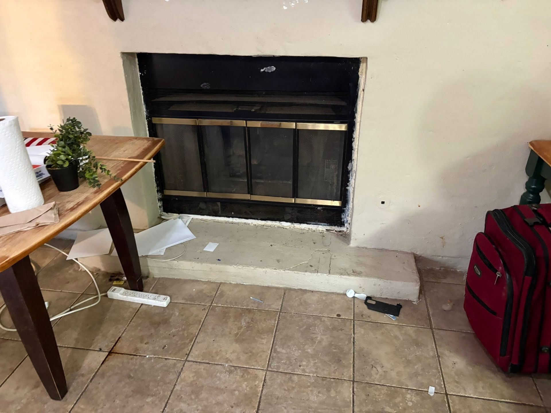 Empty fireplace with a small table, plant, and red suitcase in a tiled room.