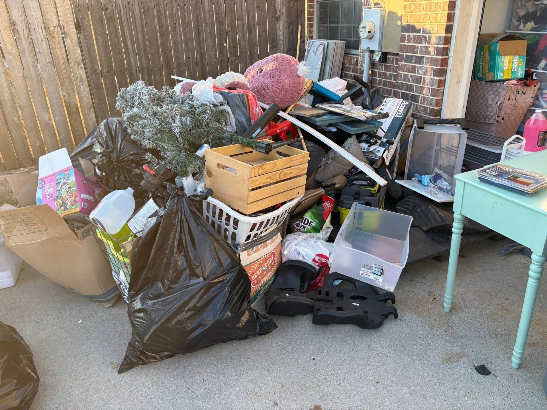 Outdoor cluttered area with bags, boxes, tools, and scattered debris beside a fence and table