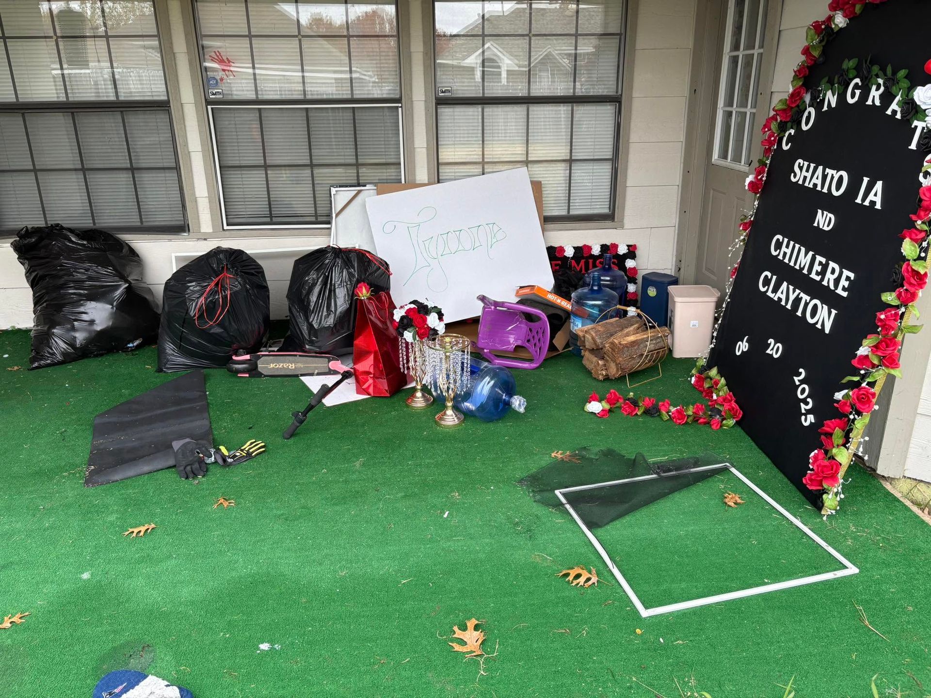 Memorial display with flowers, signs, candles, and balloons on artificial grass under a windowed porch