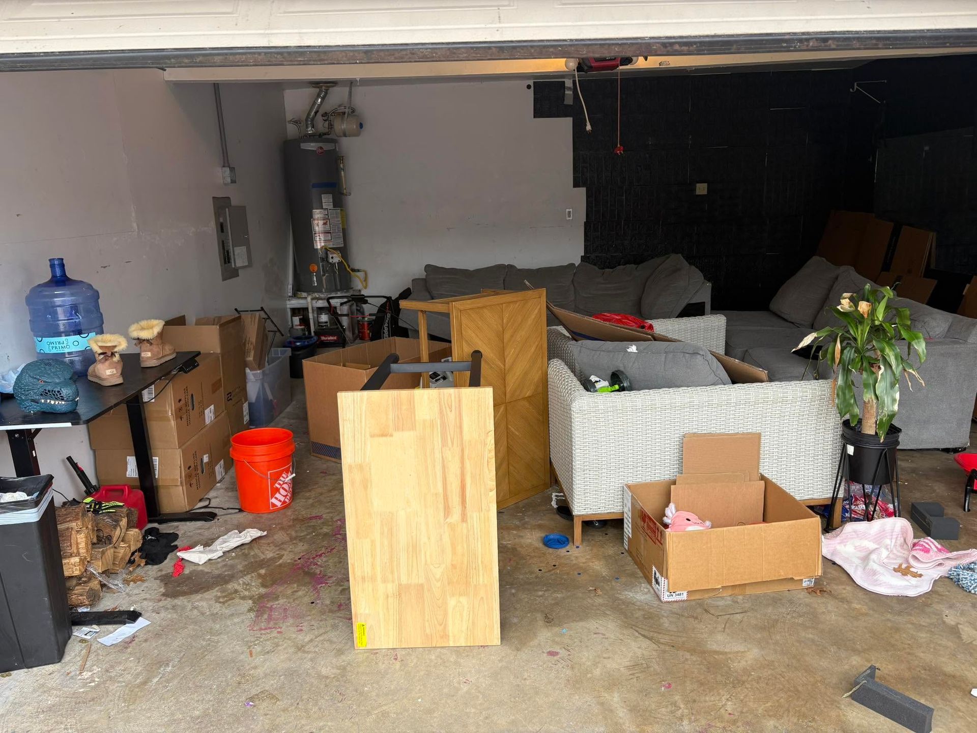 Cluttered garage with boxes, furniture, a wooden panel, and household items scattered around.