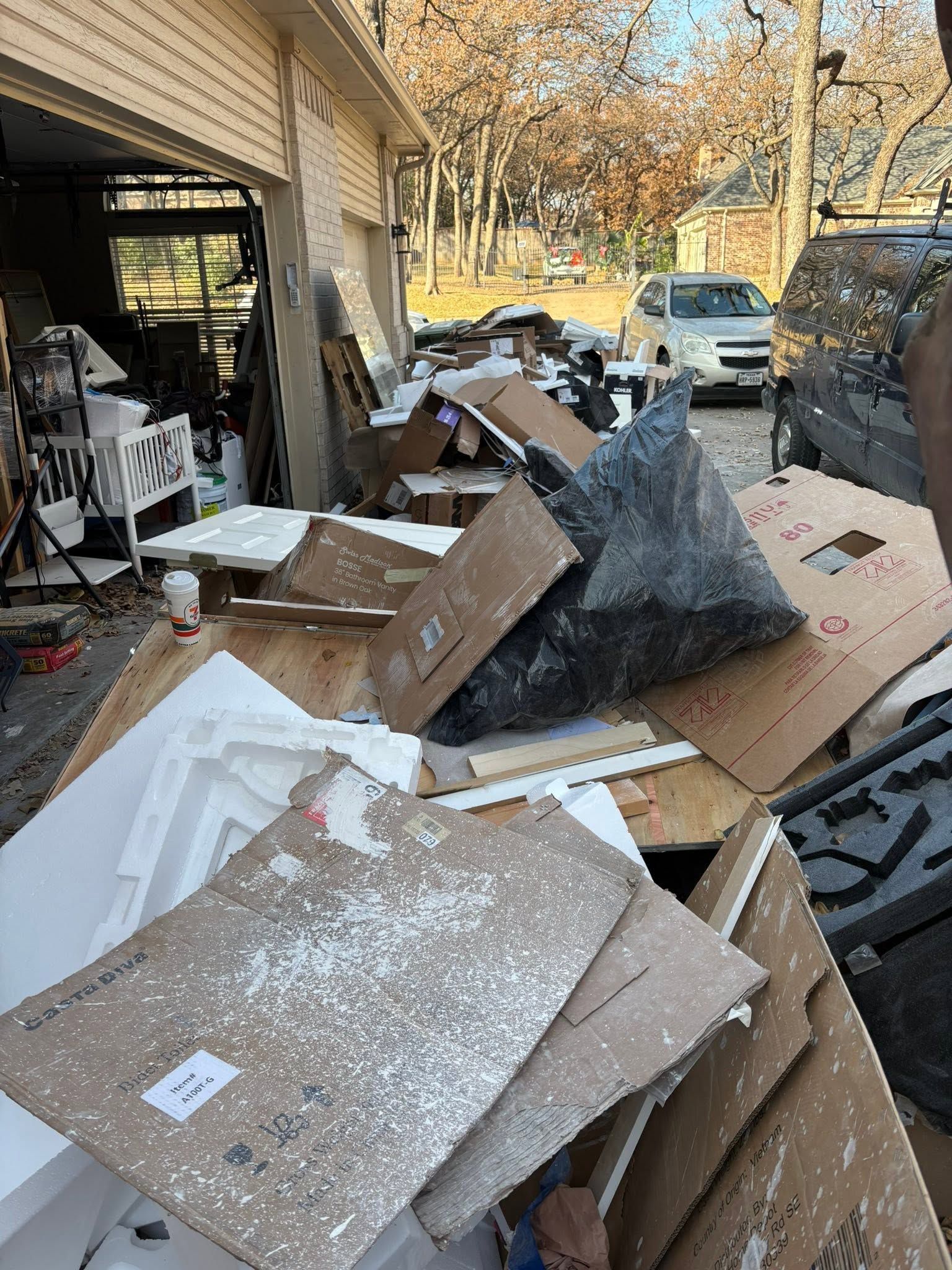 Pile of broken boxes and packing debris cluttering a garage driveway beside a parked car.