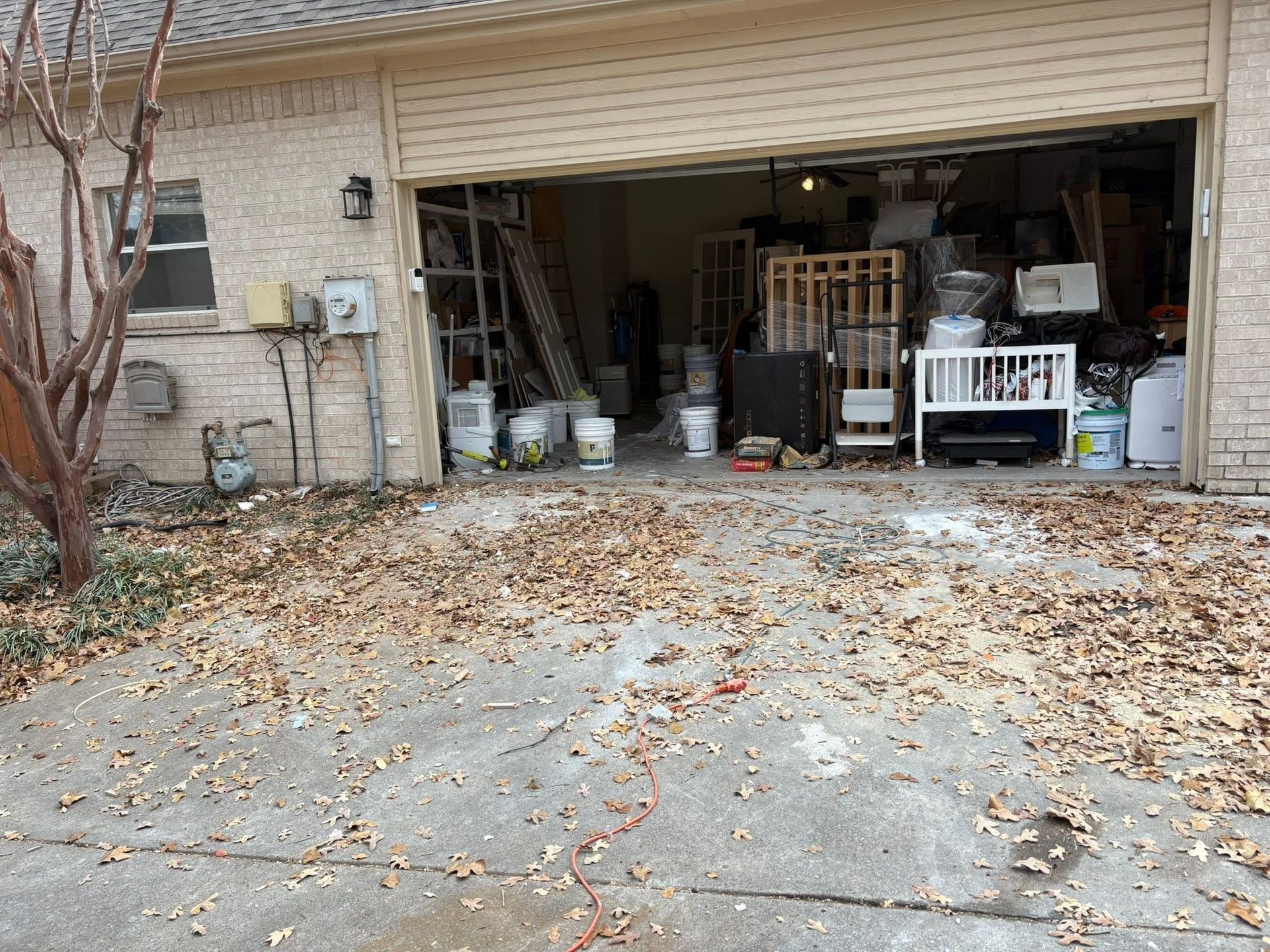 Open garage with cluttered storage and leaf-covered driveway in front of a beige house