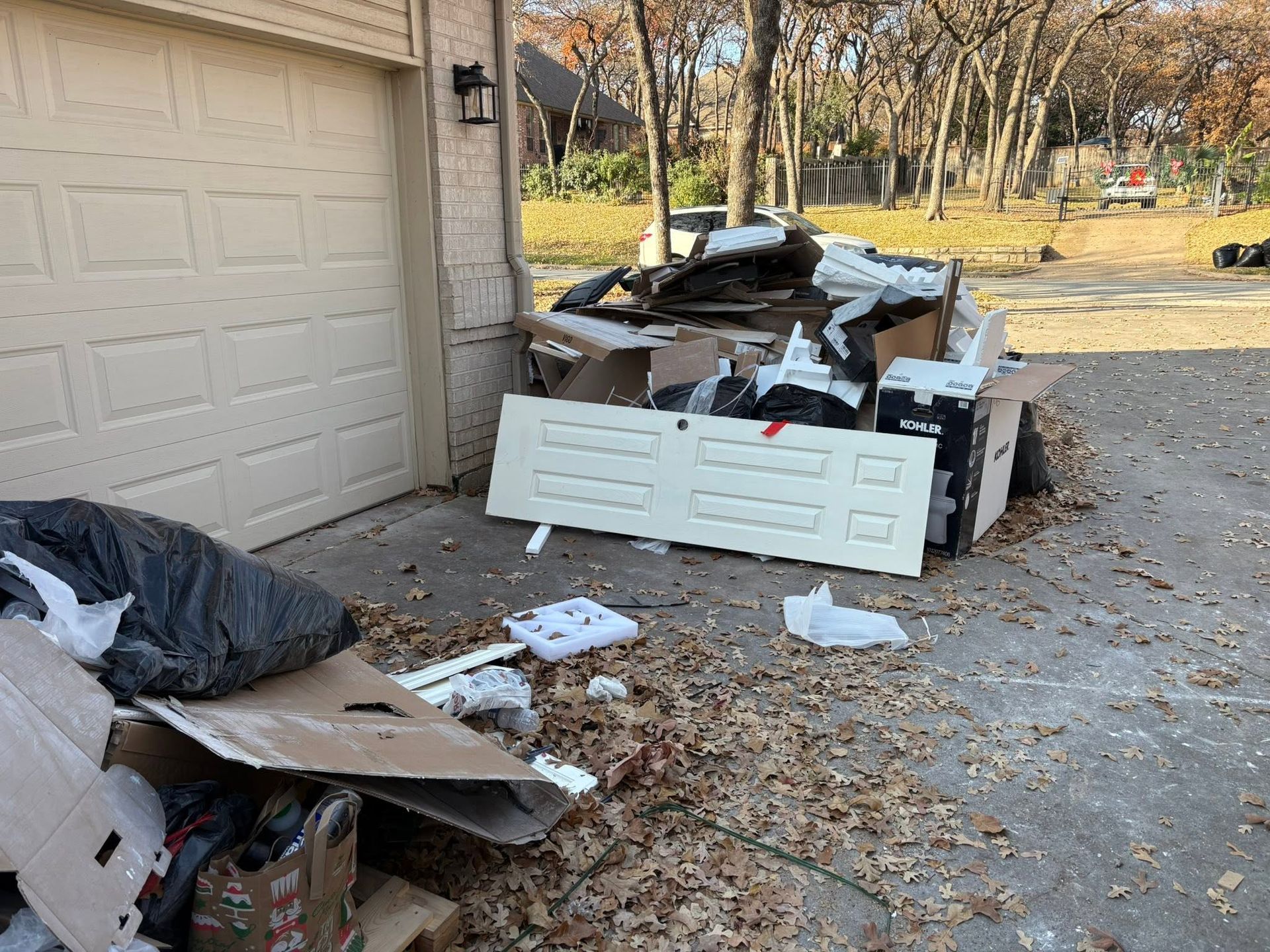 Overflowing curbside trash and debris piled beside a garage driveway on a leaf-covered ground