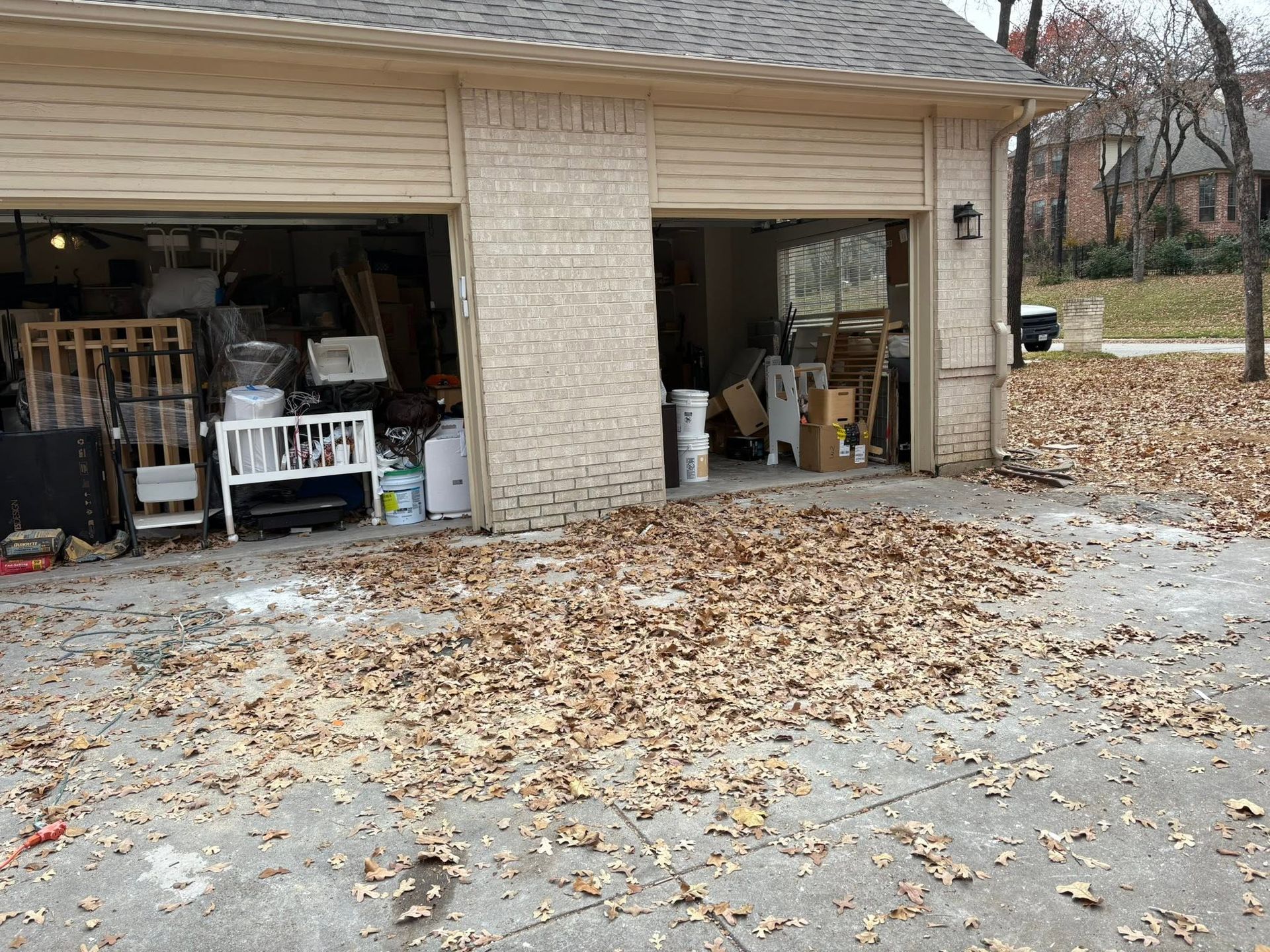 Garage with both doors open, cluttered with stored items, and leaves scattered on the driveway.
