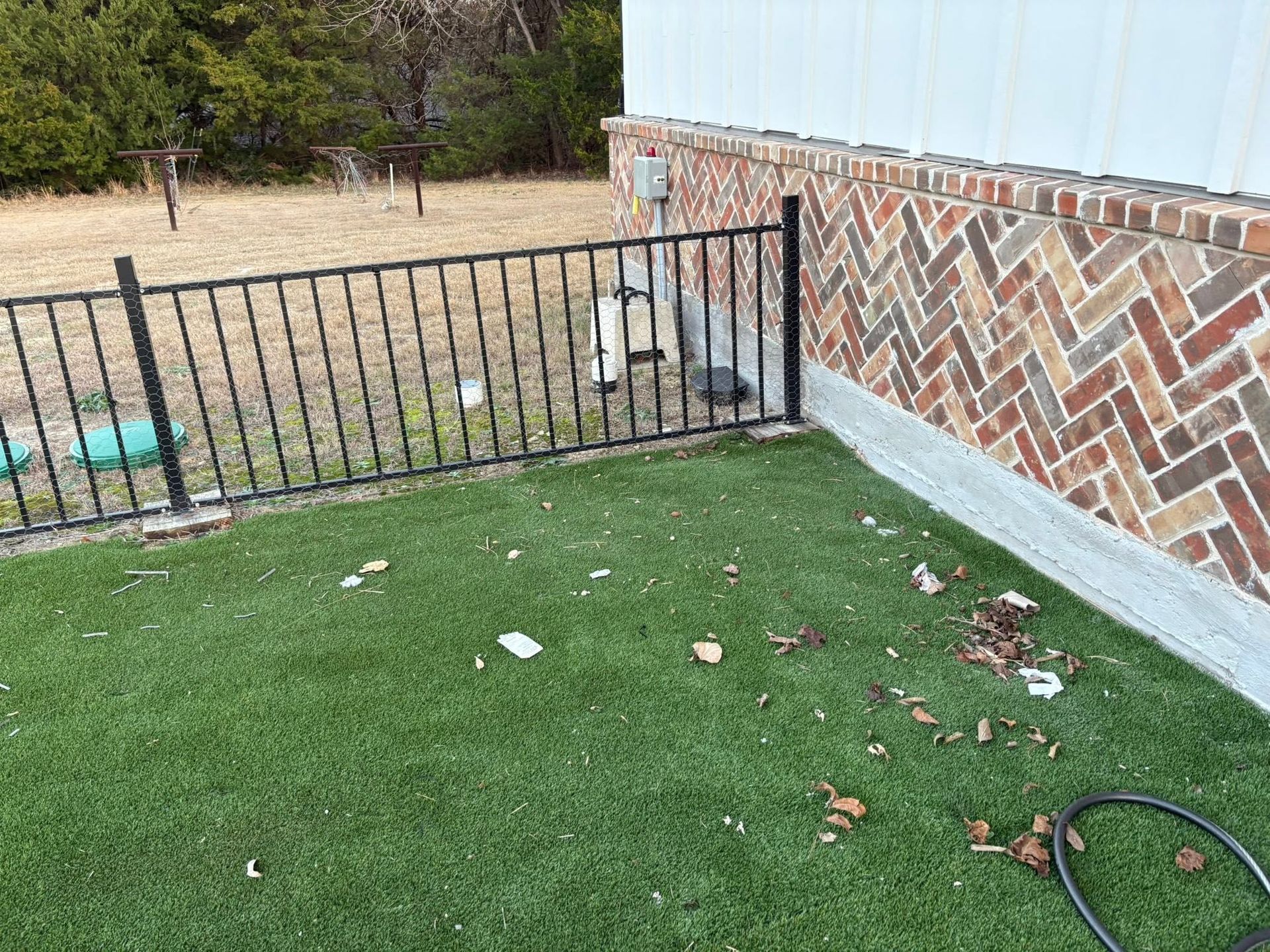 Backyard with black metal fence, green lawn, and brick house wall with a hose on the ground