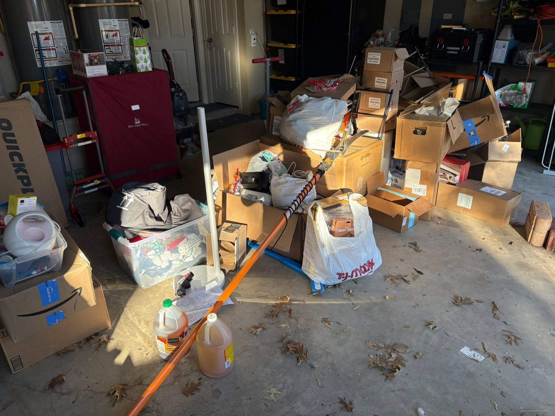 Cluttered garage with scattered boxes, bags, and household items in bright sunlight.