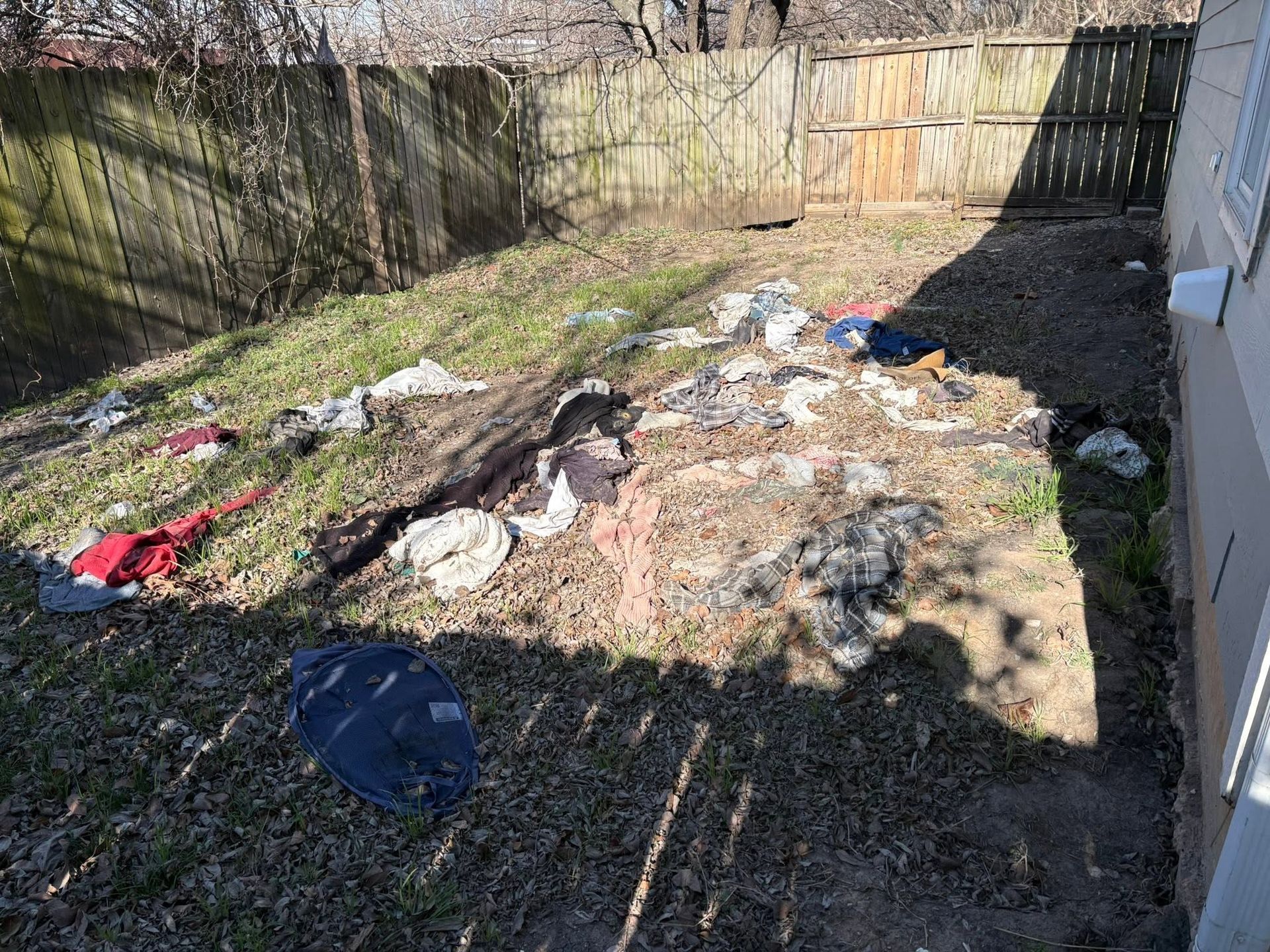 Scattered trash and clothing in a fenced backyard with dirt and grass patches.