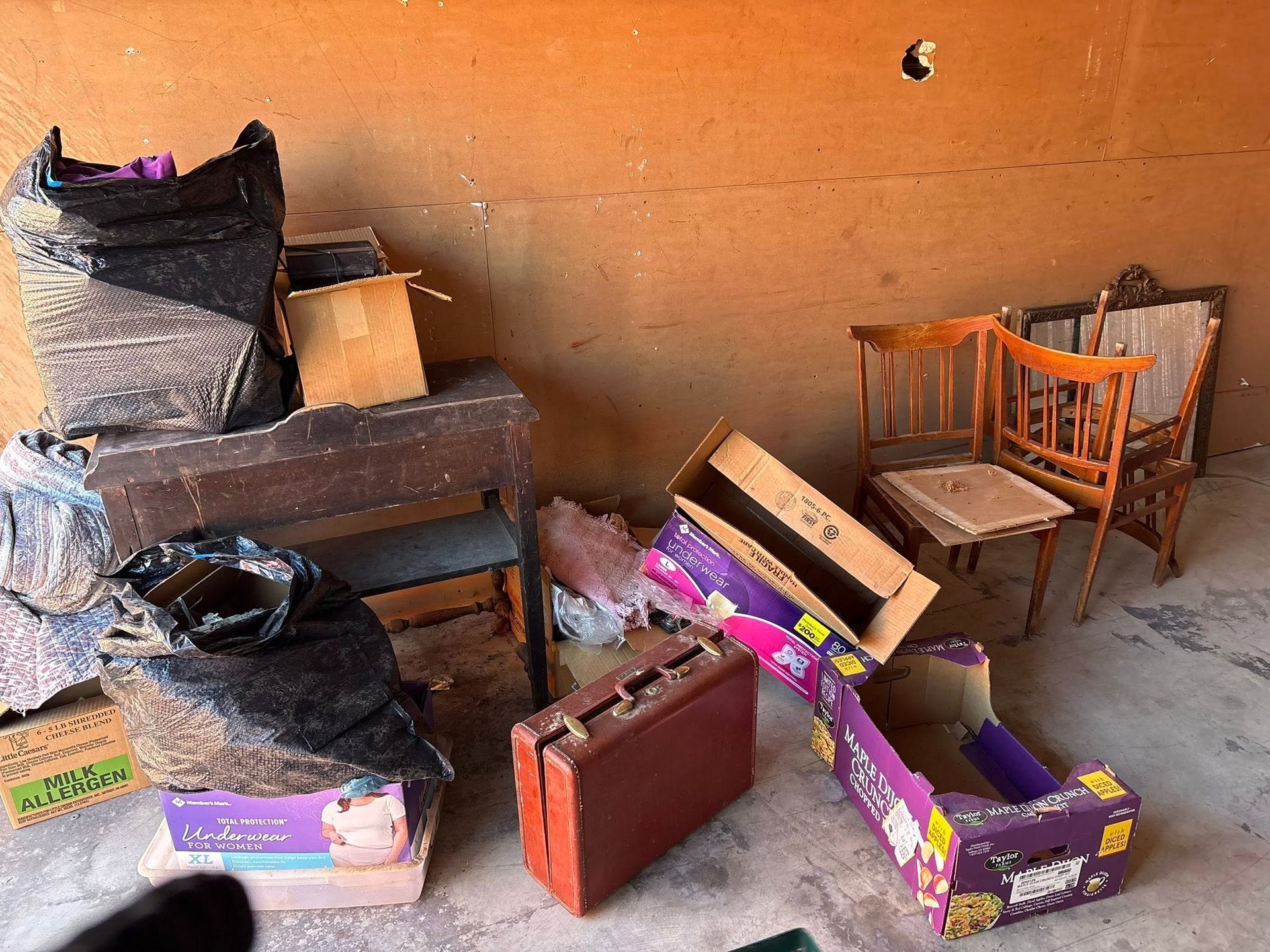 Scattered luggage, cardboard boxes, and two wooden chairs against an orange wall in a cluttered room