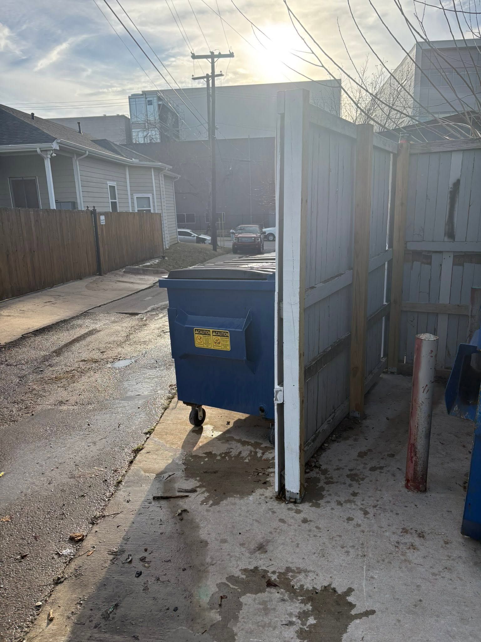 Blue dumpster by open white gate on a wet driveway in an alley, with houses and utility poles in the background