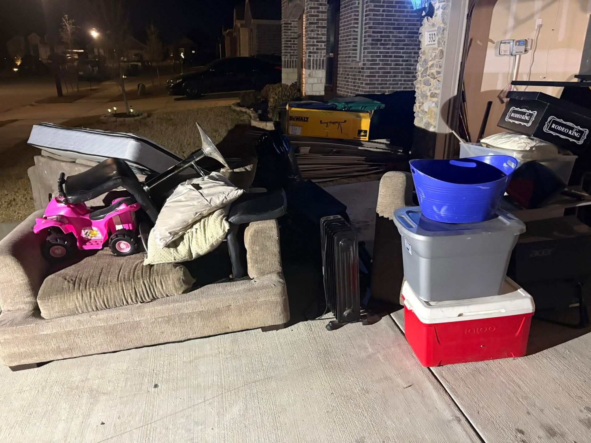 Nighttime porch with stacked furniture, storage bins, a blue bucket, and a pink toy car on a concrete driveway