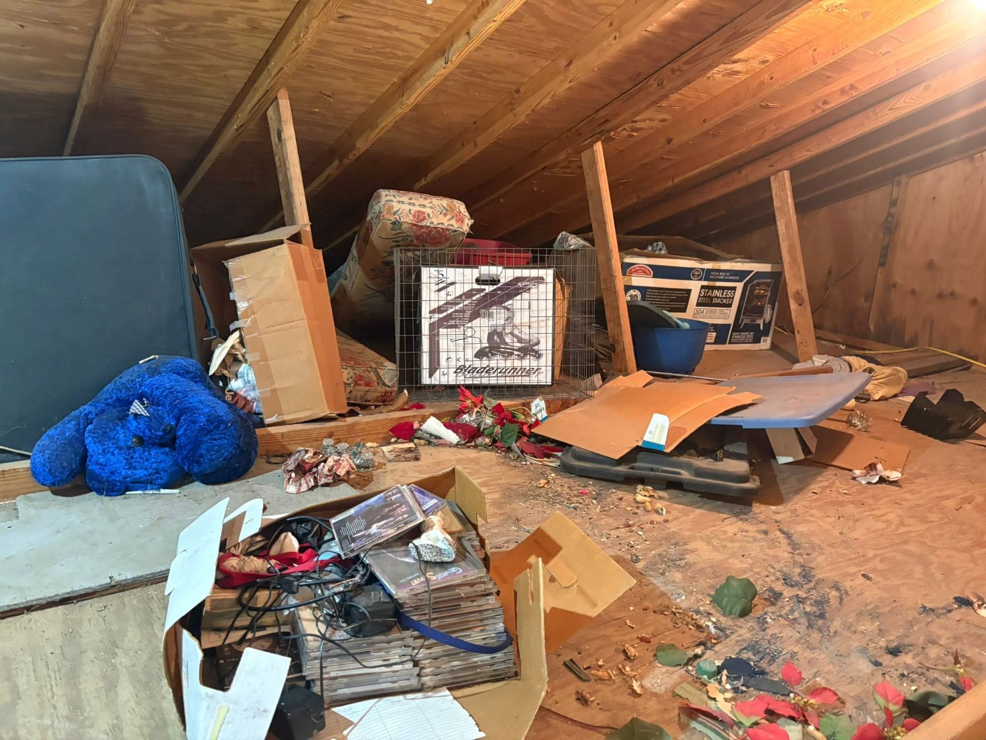 Cluttered attic with scattered debris, boxes, papers, and a blue stuffed toy under a slanted wooden roof