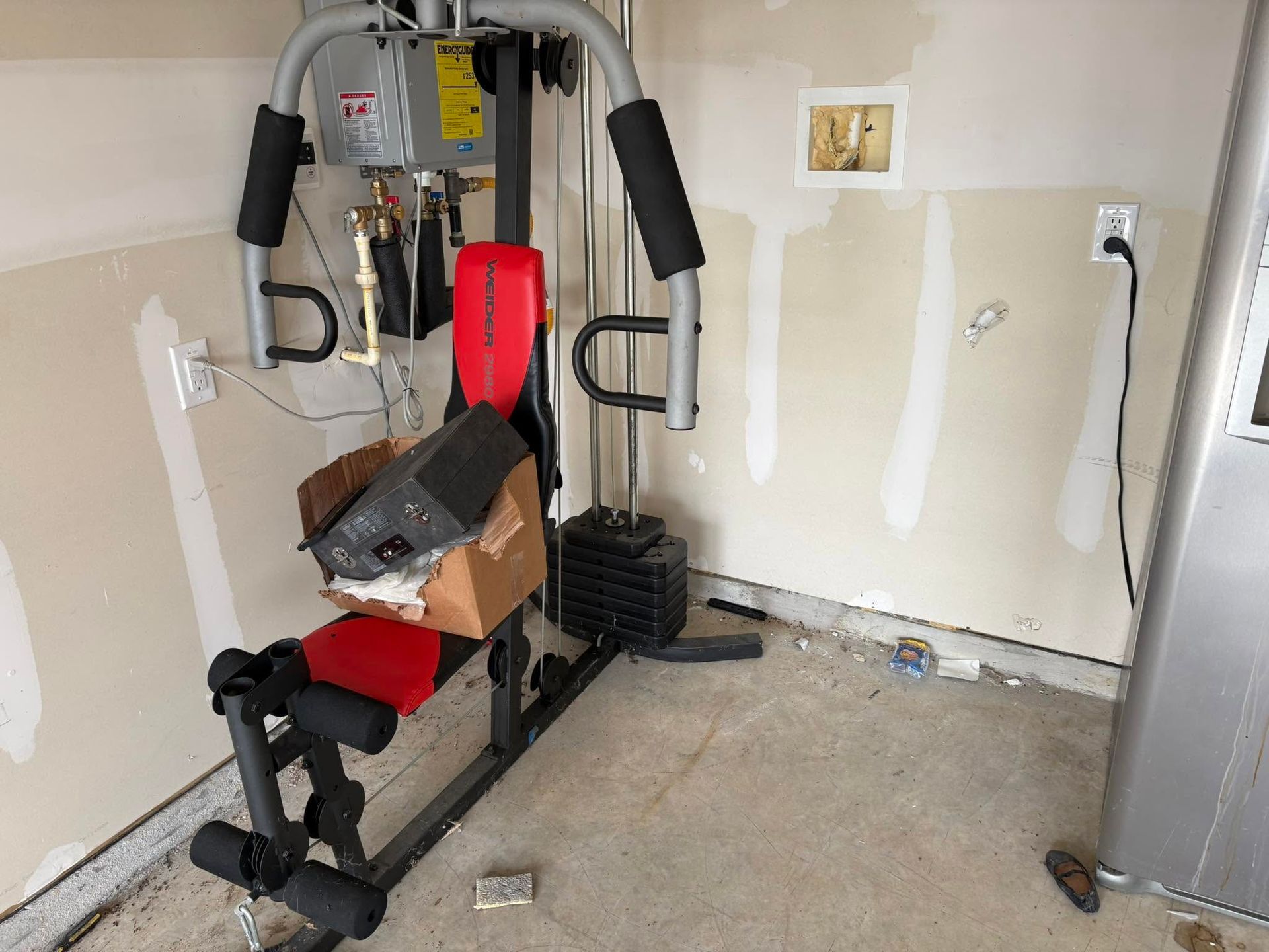Red weight machine with boxes in a garage next to a water heater and unfinished walls