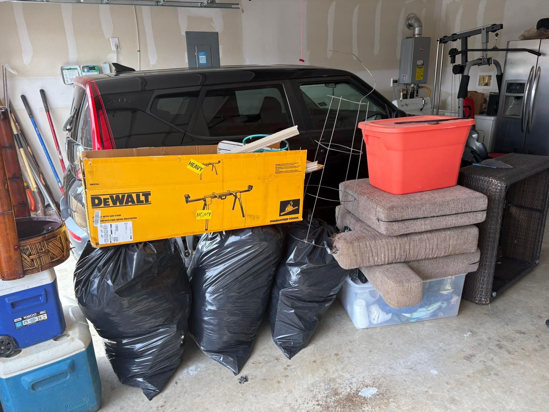 Garage cluttered with bags, stacked bins, and a yellow DEWALT box in front of a black vehicle.