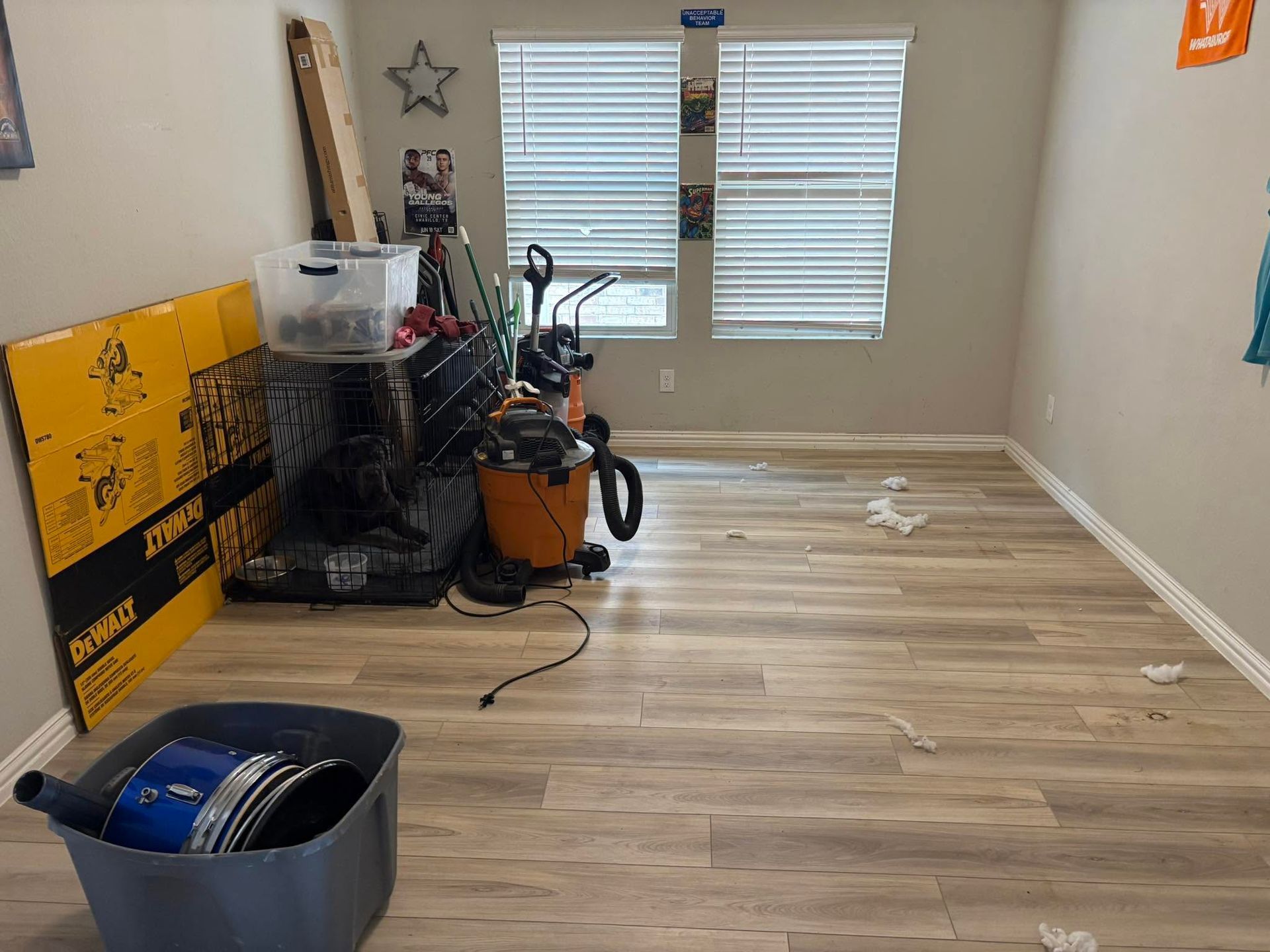 Empty room with light wood floor, cleaning equipment, and a plastic bin near windows.