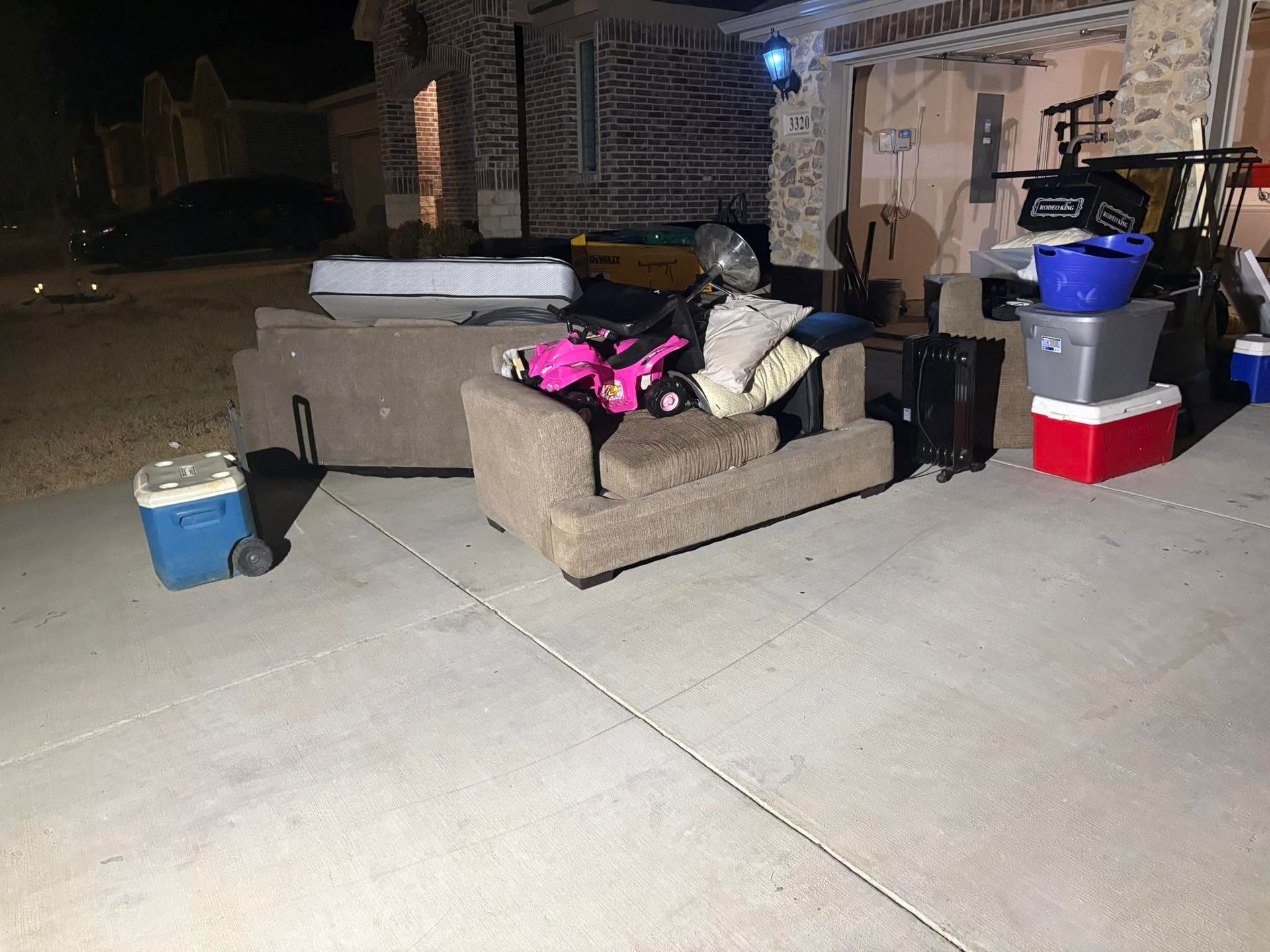 A couch on a driveway piled with bags and bins, with a cooler and boxes near a house at night.