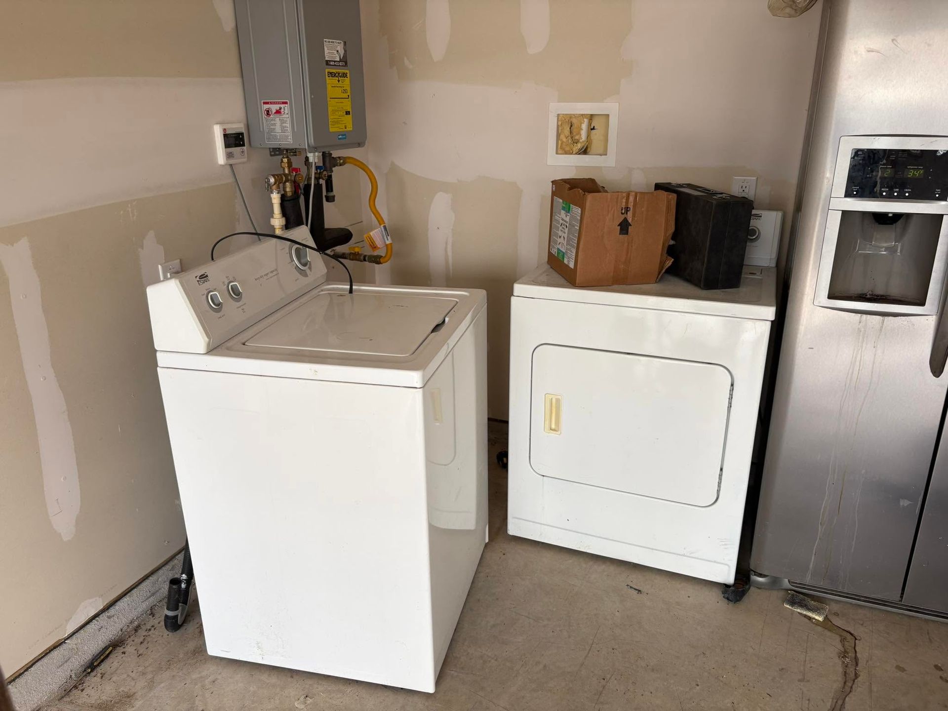Laundry room with a white washing machine, dryer, and stainless refrigerator against unfinished walls.