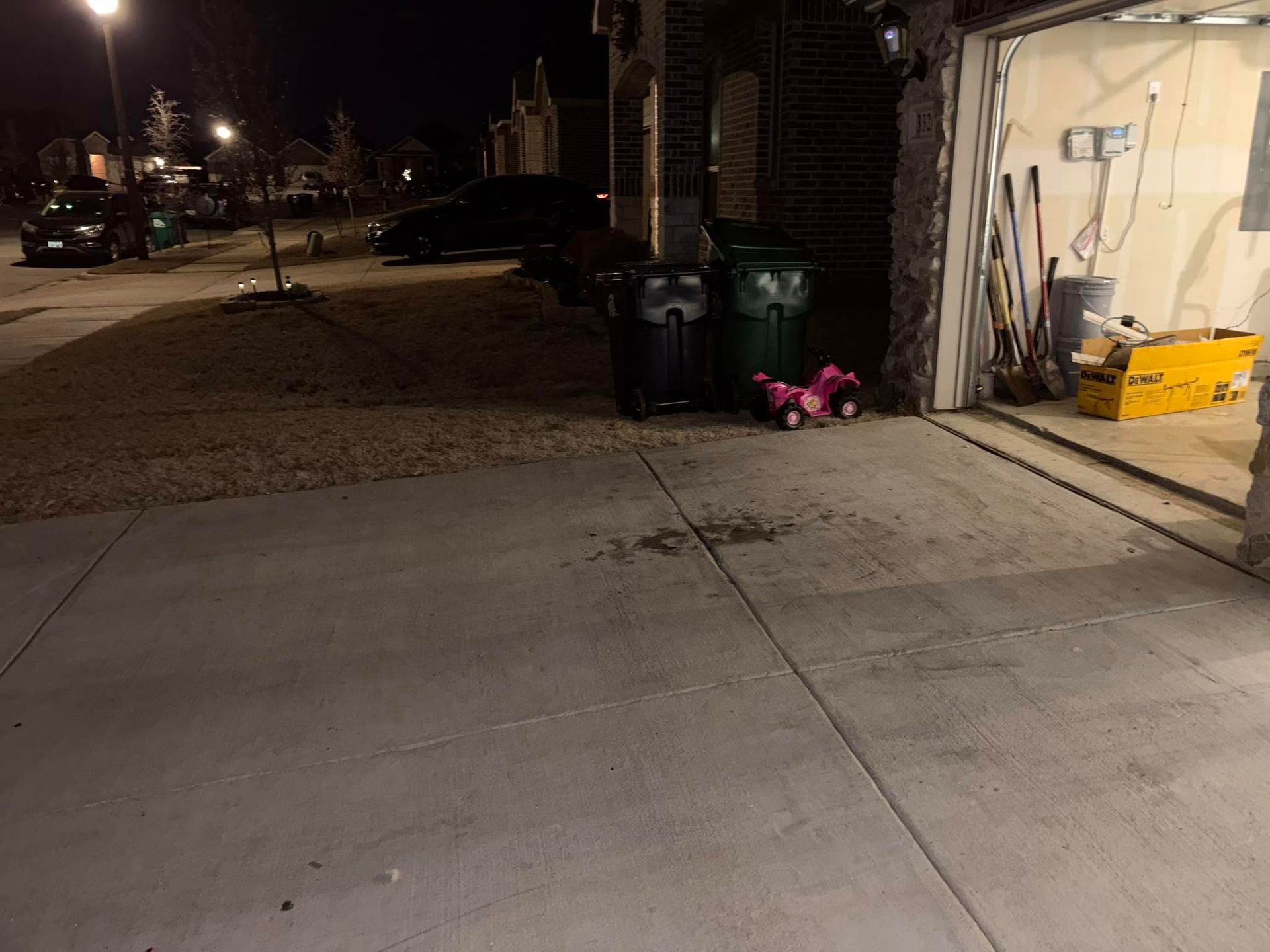 Nighttime sidewalk beside a garage, with trash bins and a small pink toy near the open door.