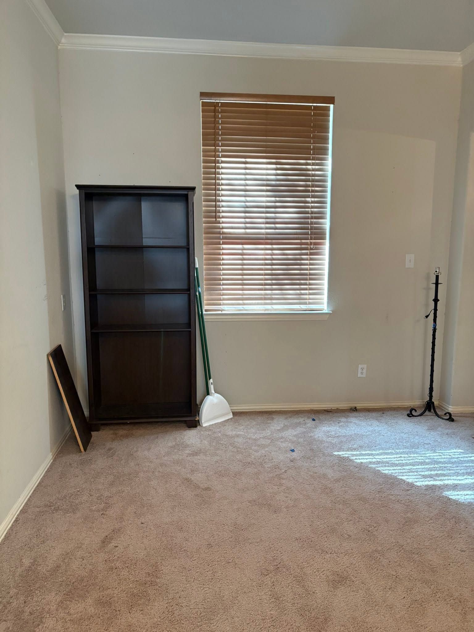 Empty beige carpeted room with window blinds, dark shelf, and cleaning tools against the walls