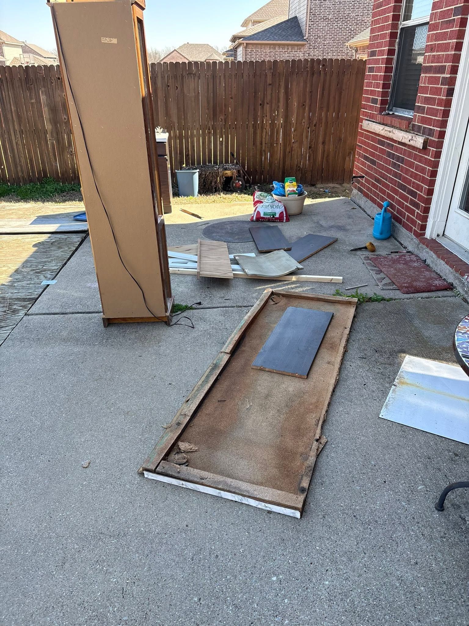 Backyard patio with broken concrete slabs, a fallen wooden panel, and tools near a brick house and fence