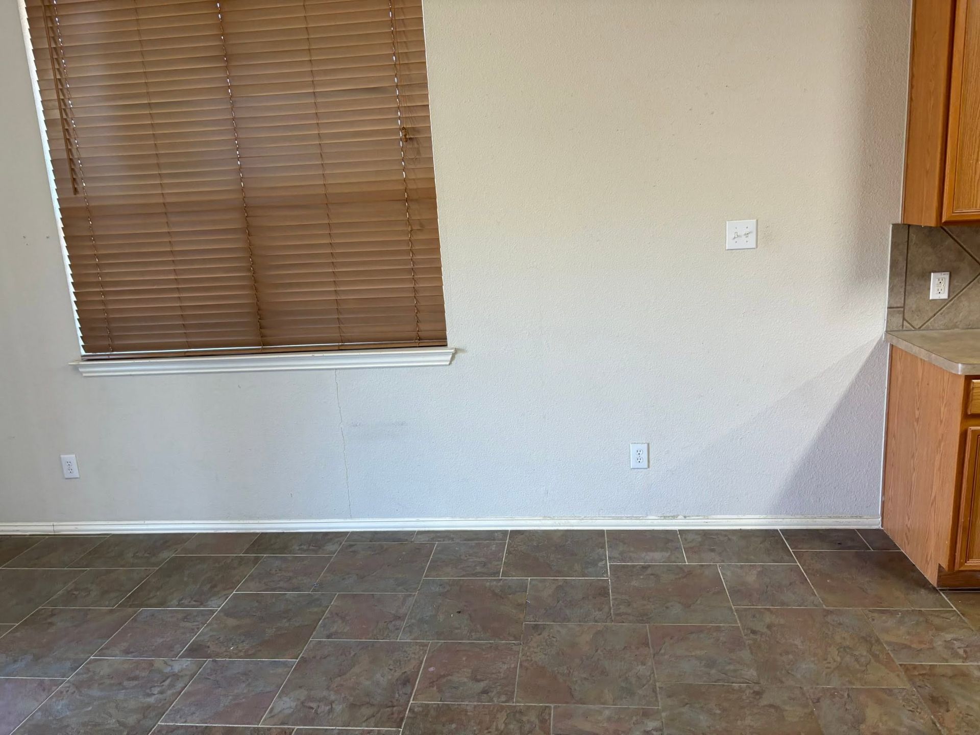 Empty room with tiled floor, white walls, window blinds, and kitchen cabinets on the right