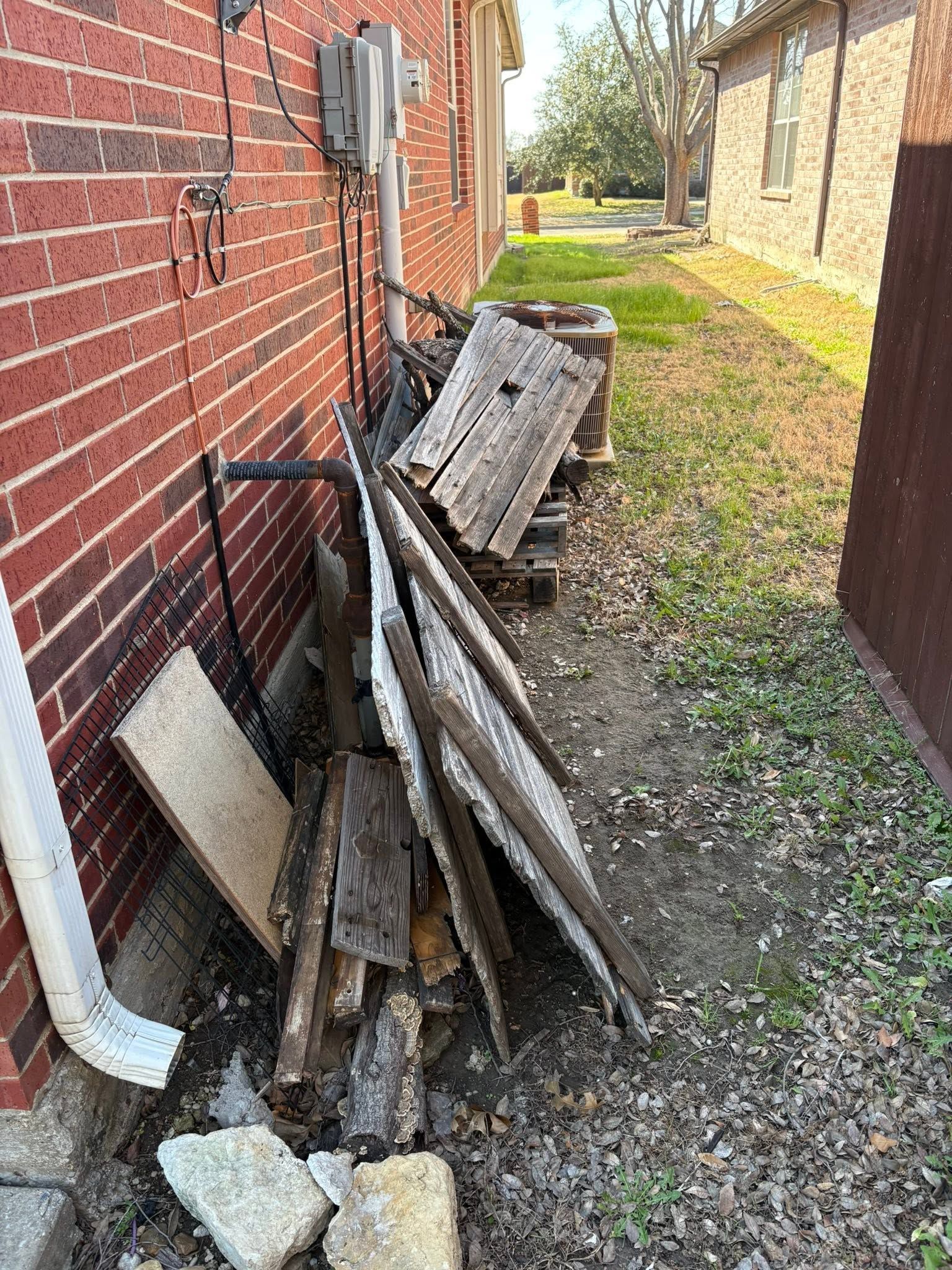 Piles of old lumber and debris stacked beside a red brick house along a narrow grassy alley