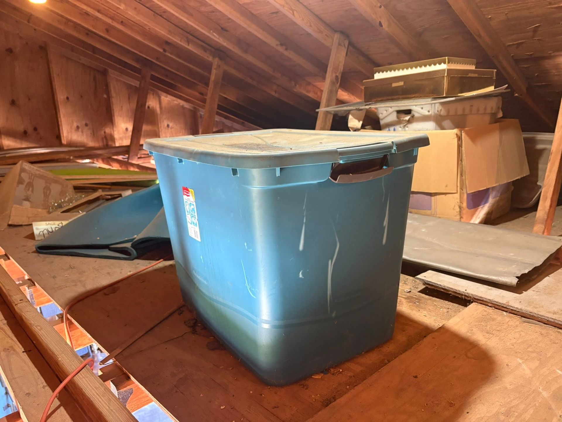 Blue storage bin in a cluttered attic with boxes and wooden boards.