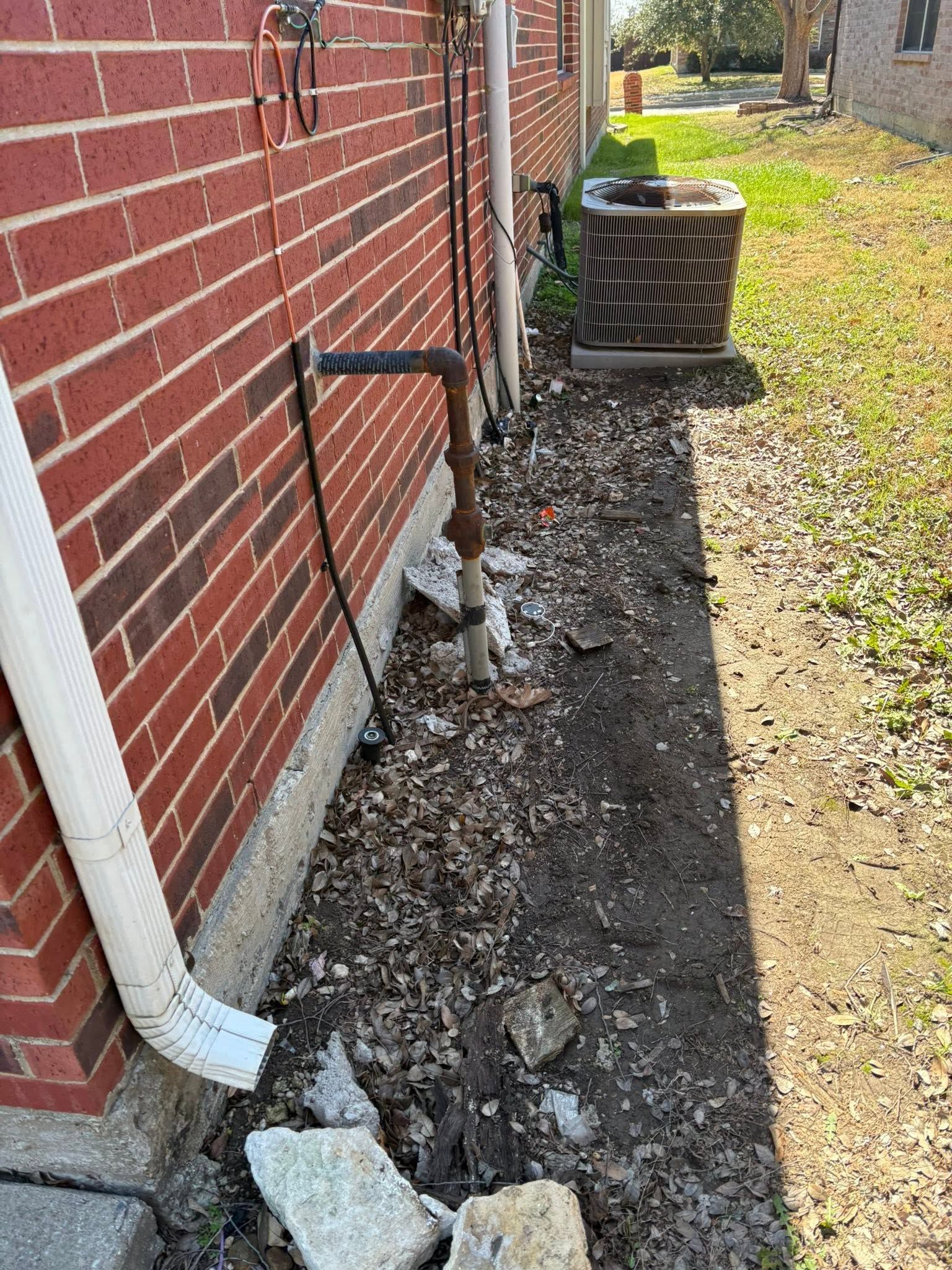 Narrow gravel strip beside red brick house with white downspout and black pipe near outdoor AC unit