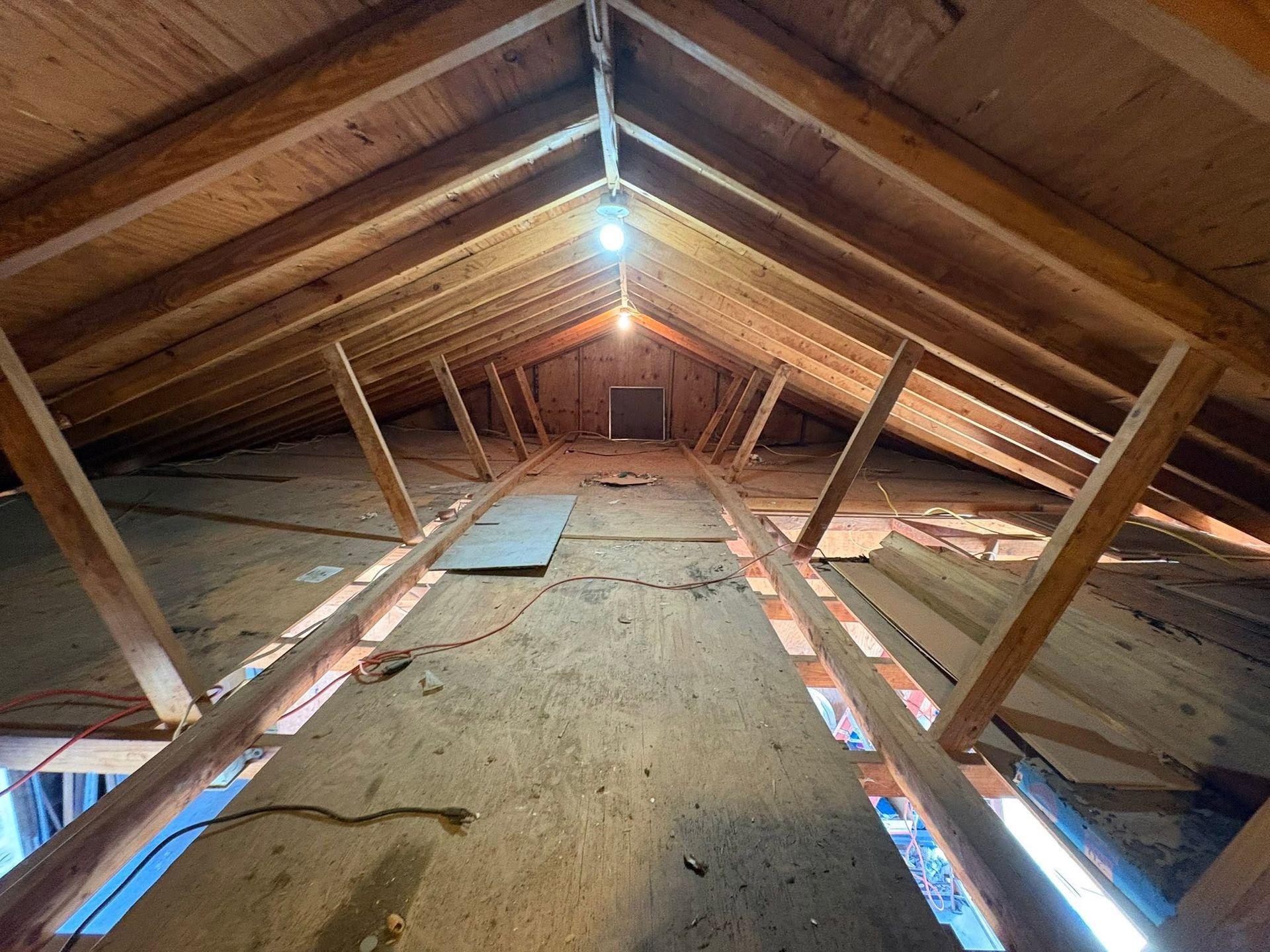 Attic interior with exposed rafters, insulation, and a single hanging light bulb, viewed from the center beam.