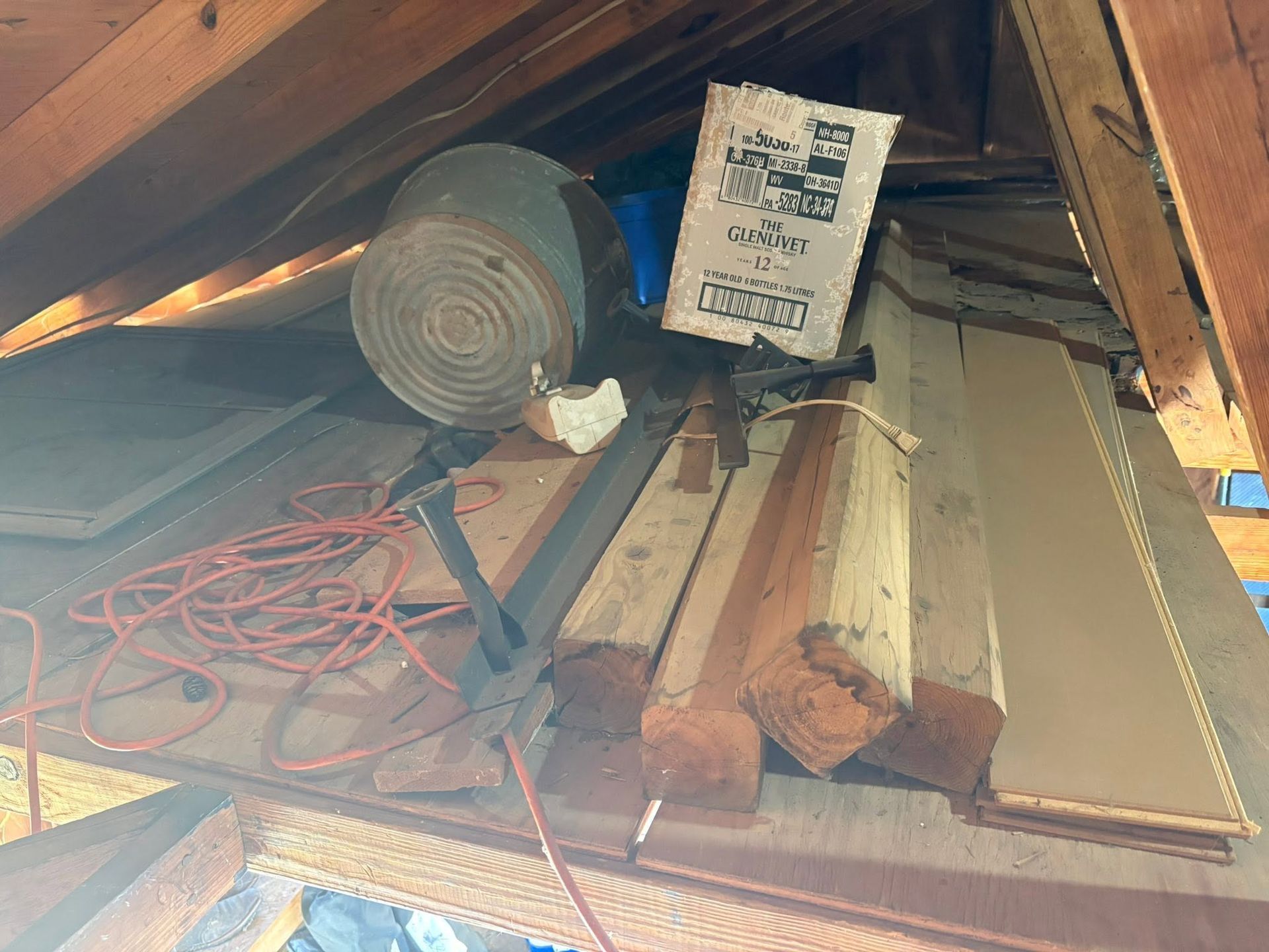 Cluttered attic with duct insulation, cardboard boxes, lumber, and coiled red extension cords.