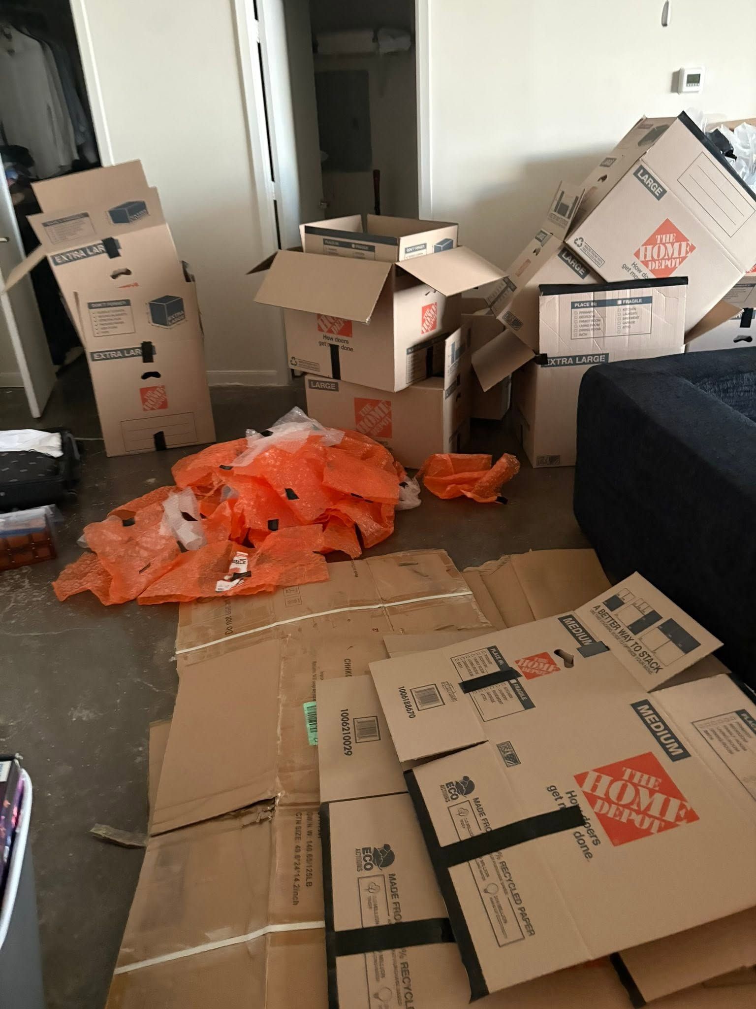 Cluttered room with stacked cardboard boxes and orange packing material scattered on the floor.