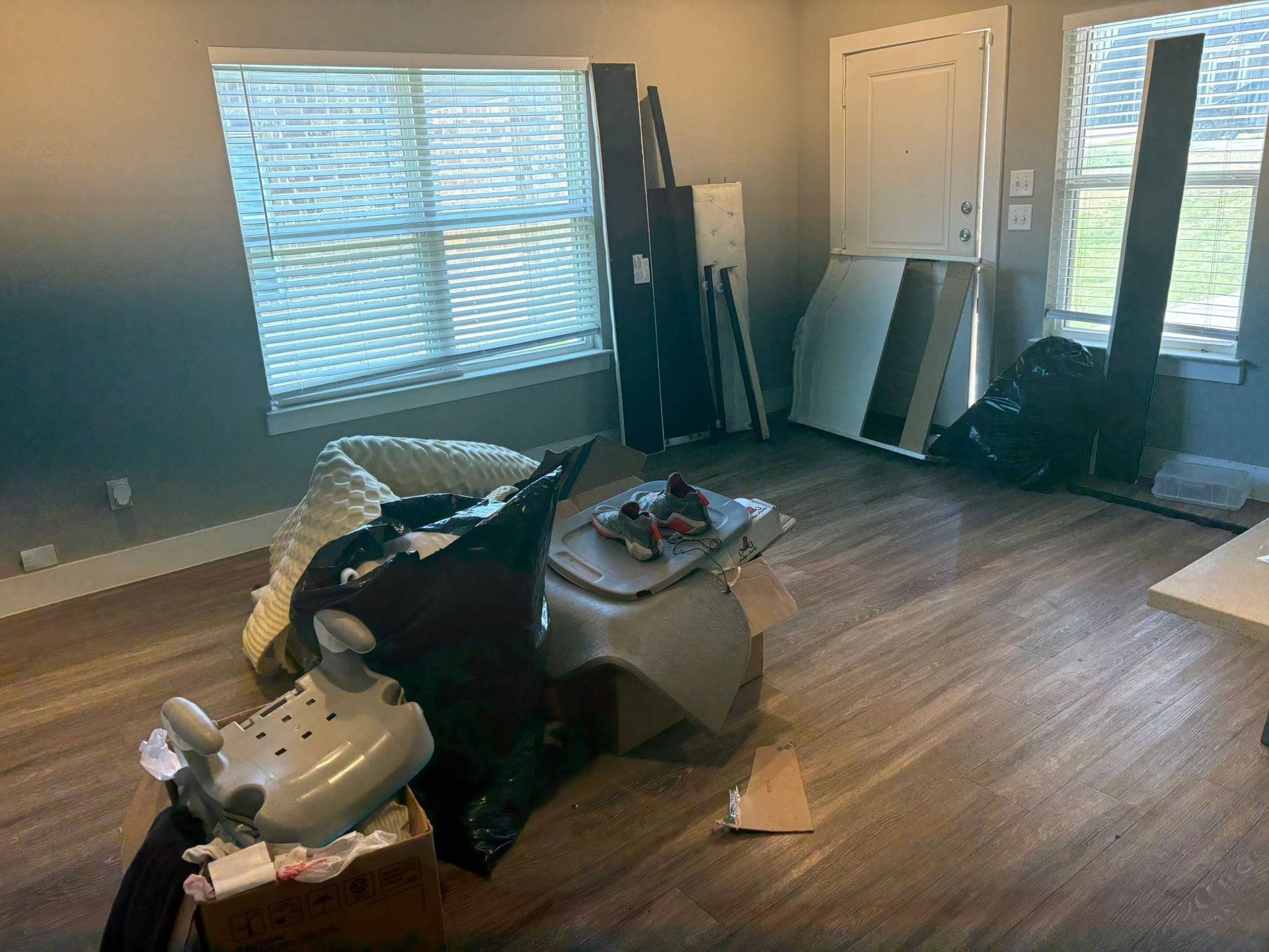 Cluttered living room with broken furniture, scattered debris, and wood flooring near a window and door