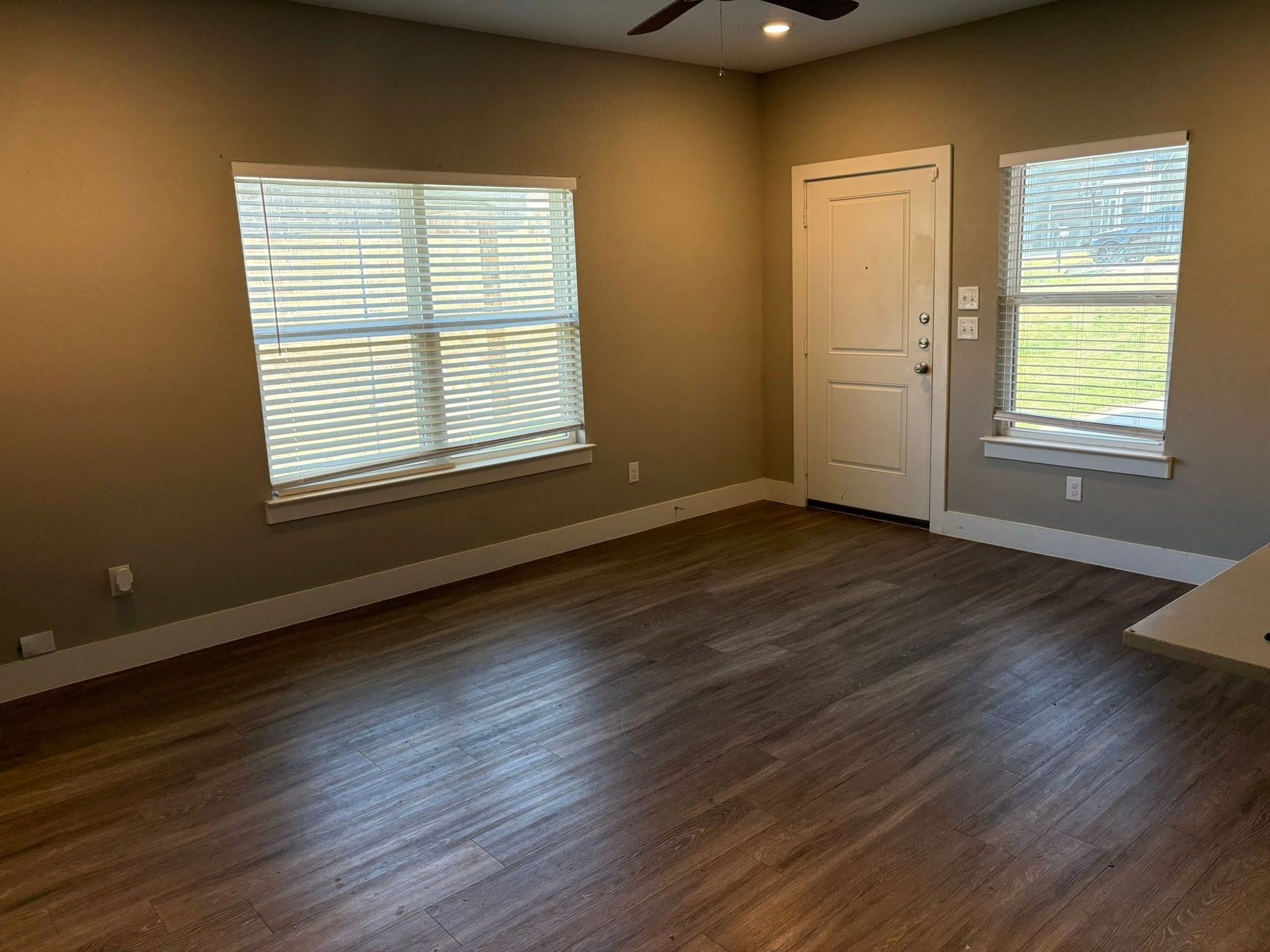Empty beige room with dark wood floors, two windows, and a white door leading outside.