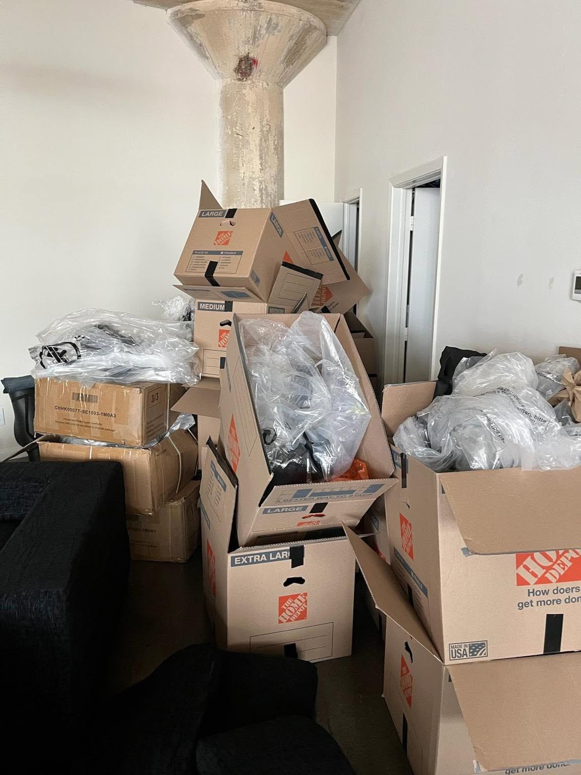 Cluttered room with stacked cardboard boxes and plastic-wrapped items piled against a wall.