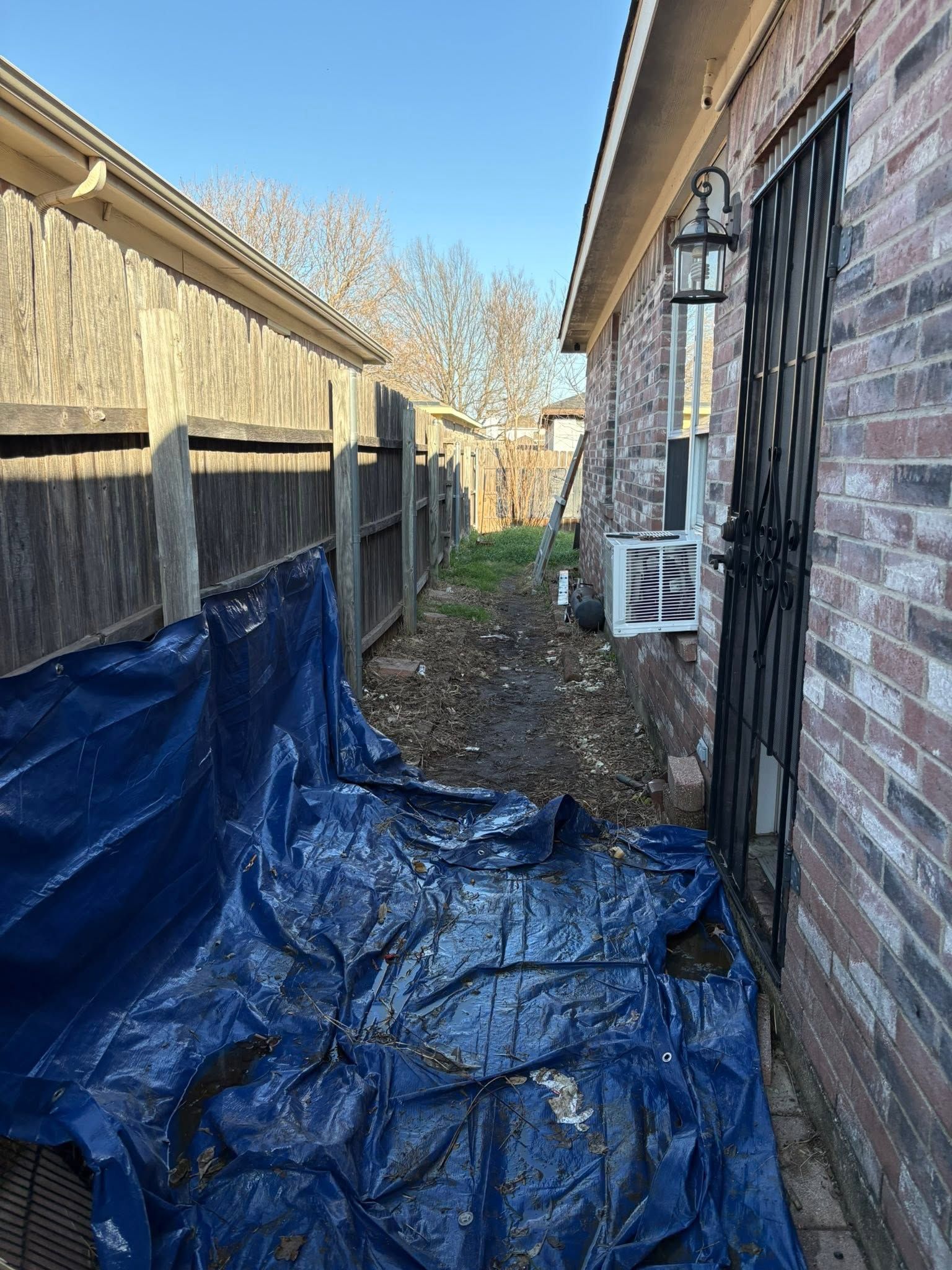 Narrow side yard with blue tarp-covered ground between a wooden fence and brick house under bright daylight